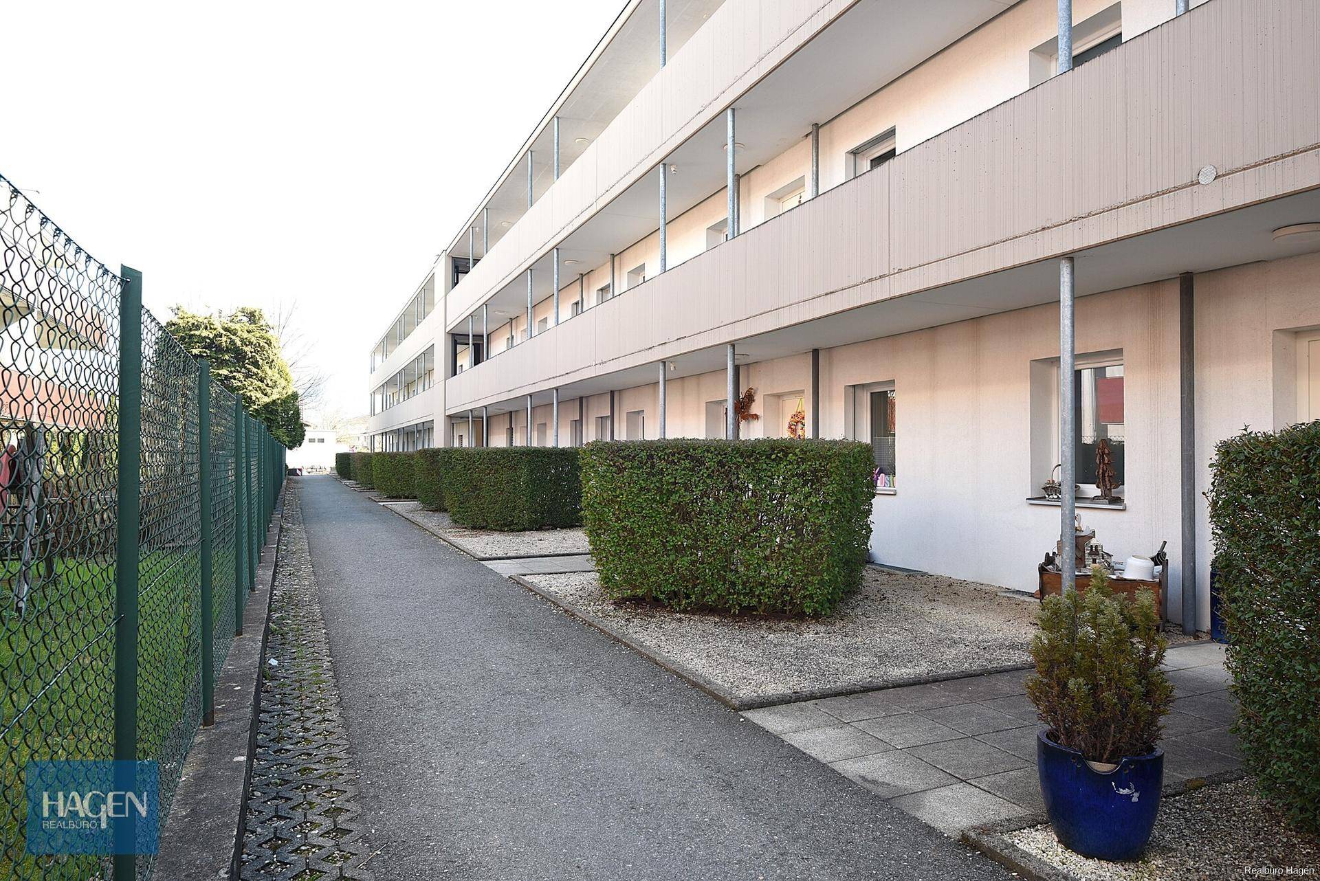 Seitenansicht eines Wohngebäudes mit einem Gehweg, Hecken und einem Zaun.