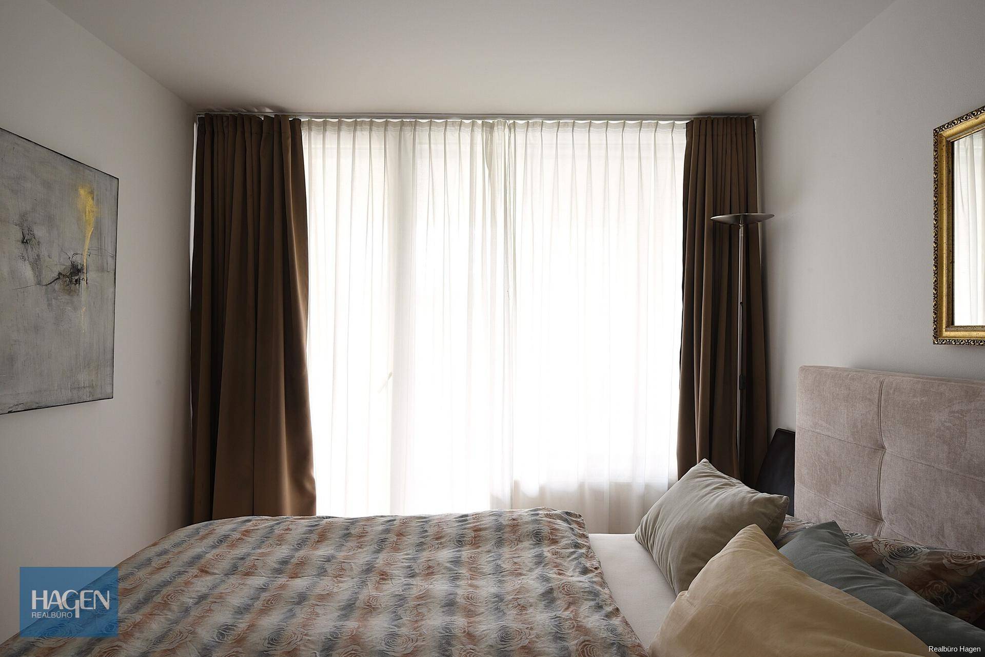 Helles Schlafzimmer mit großem Fenster, bequemen Bett und einem dekorativen Spiegel an der Wand.