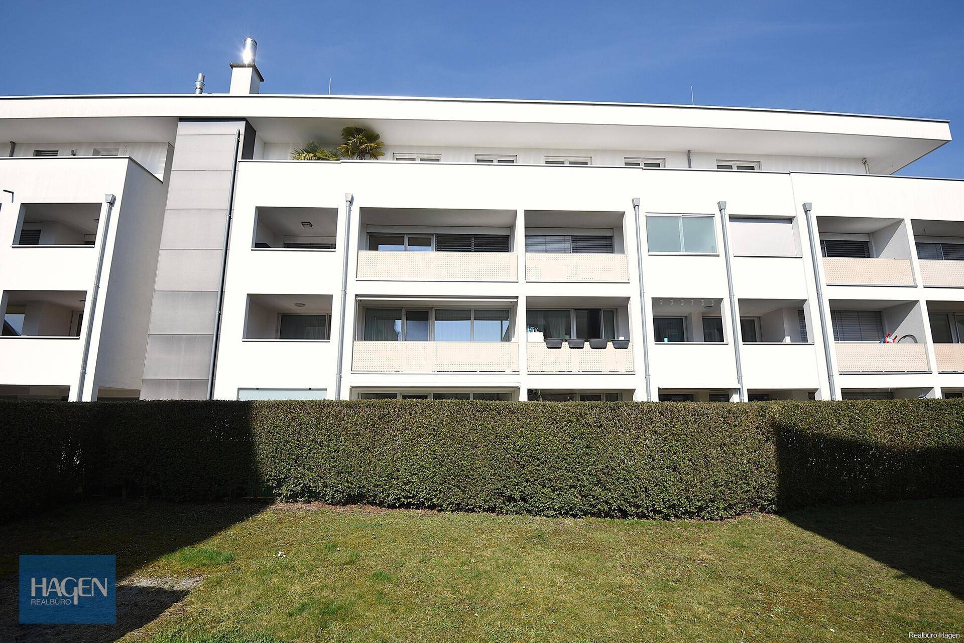 Moderne Fassade eines Wohngebäudes mit mehreren Balkonen und einer grünen Hecke im Vordergrund.
