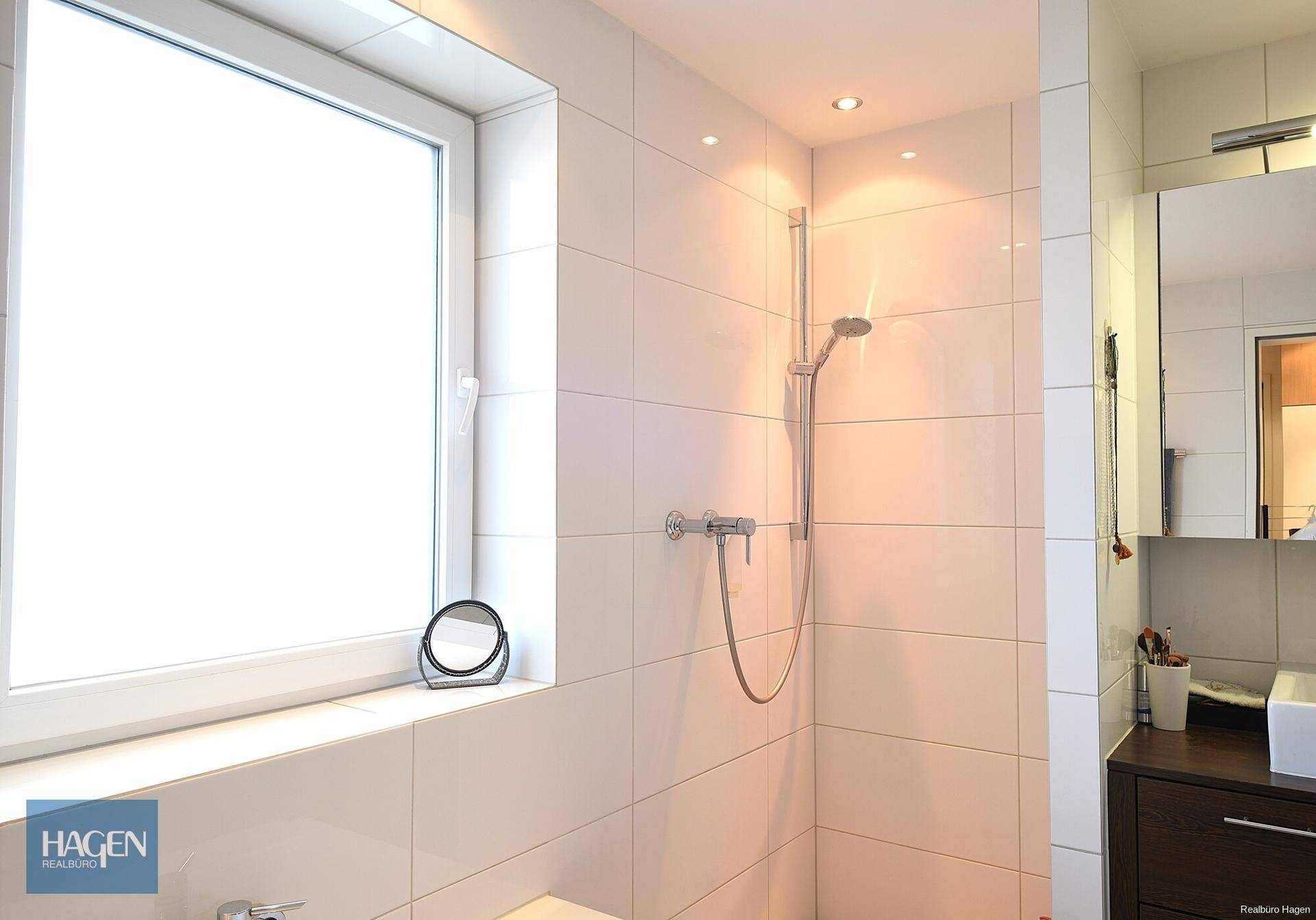 Modernes Badezimmer mit weiß gefliestem Duschbereich und einem Fenster für Tageslicht.