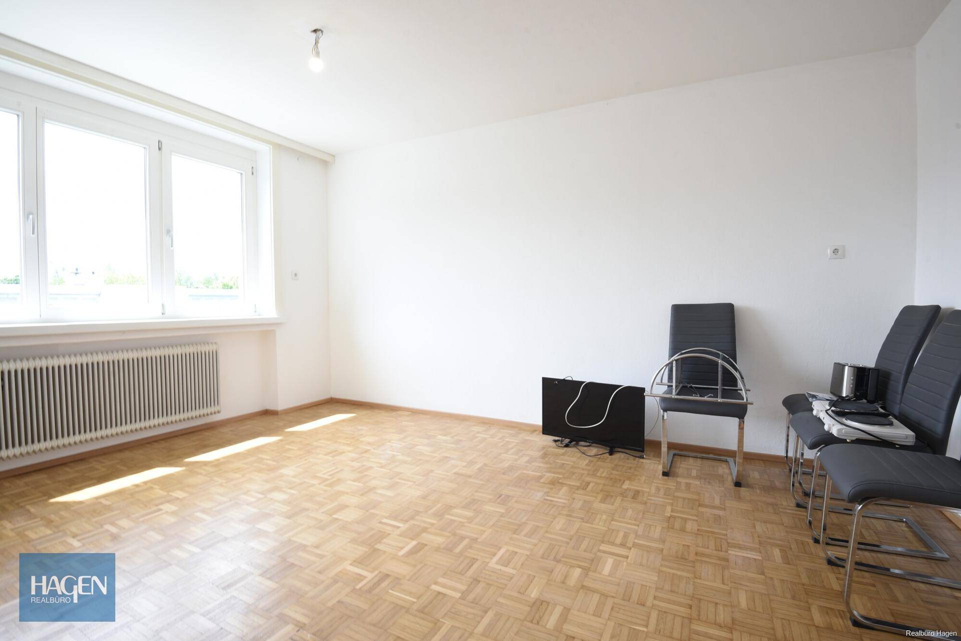 Heller Wohnbereich mit Parkettboden, großem Fenster und einfacher Möblierung, vielseitig nutzbar.