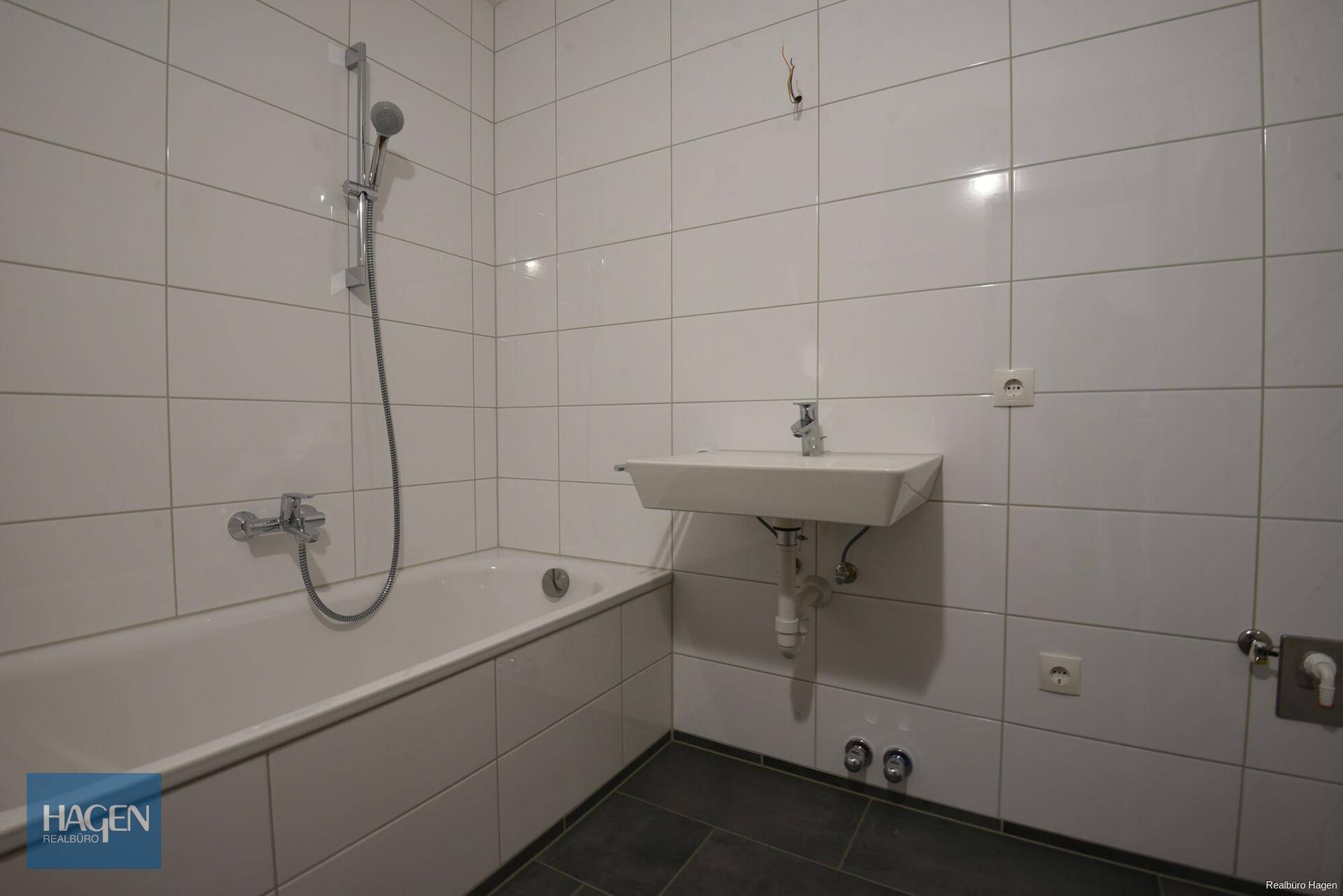 Gefliestes Badezimmer mit Badewanne, Duscharmatur und Waschbecken in schlichtem Design.