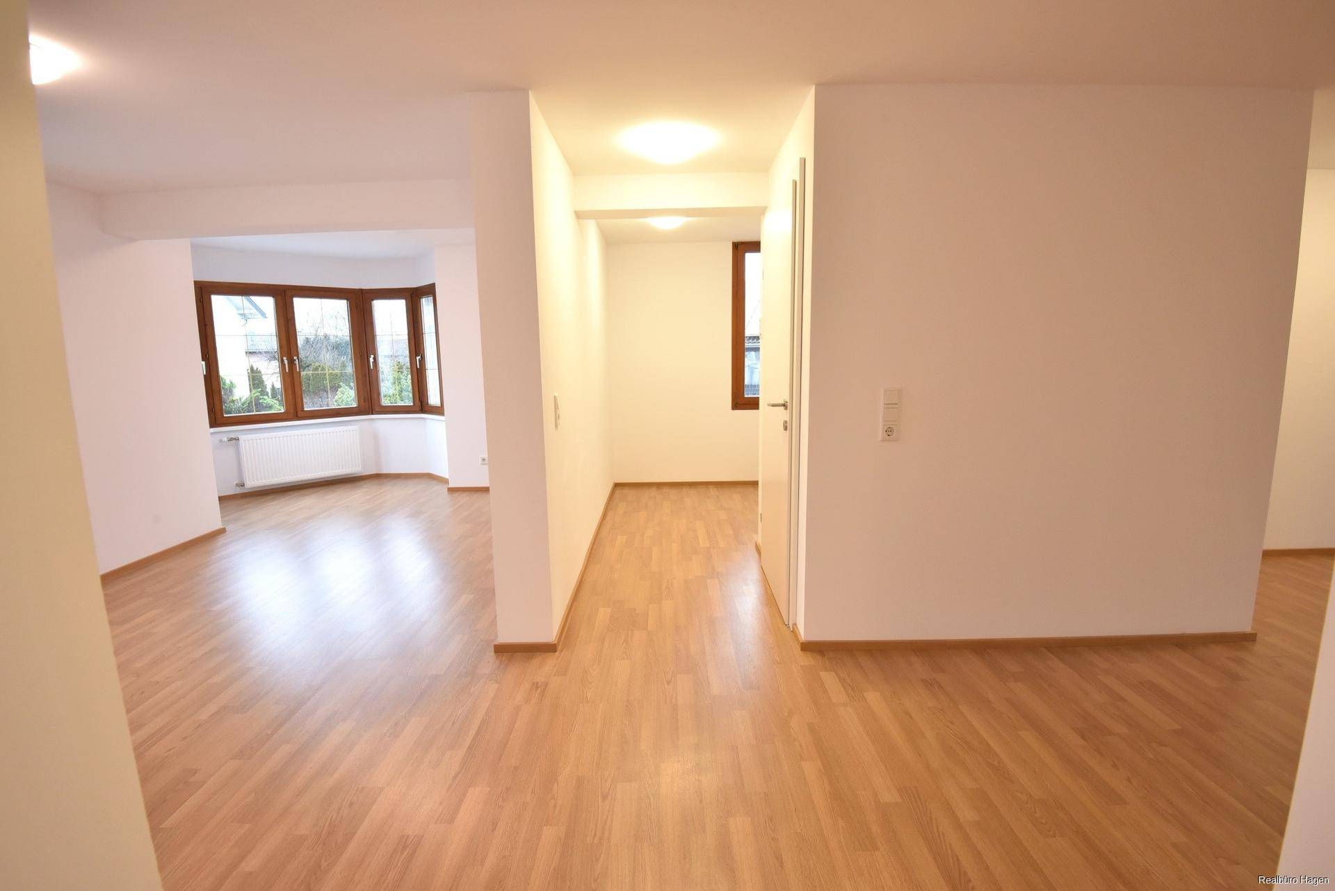 Geräumiger Wohnbereich mit Laminatboden und großen Fenstern, die viel Tageslicht hereinlassen.