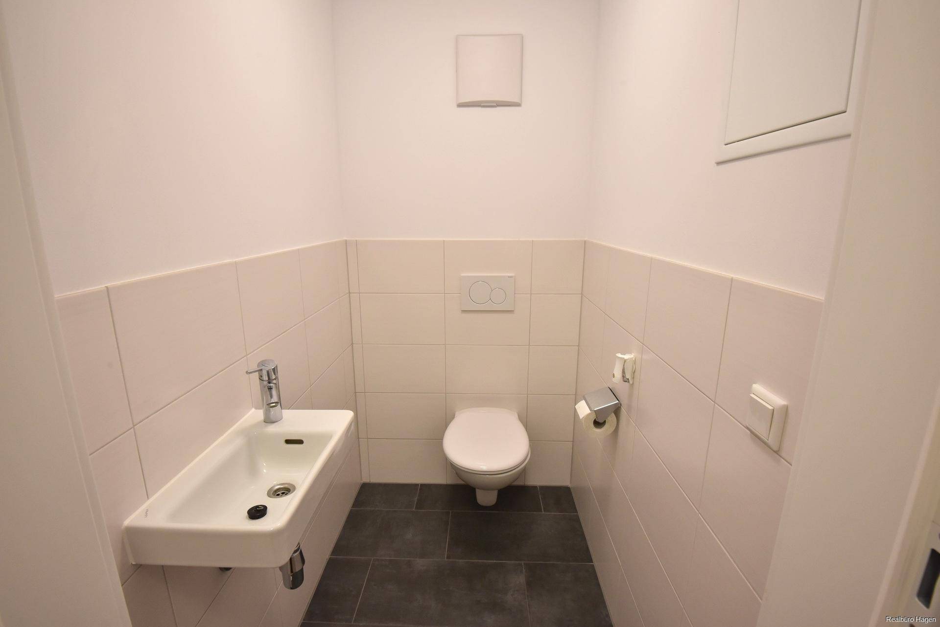 Separates Gäste-WC mit Wand-WC, kleinem Waschbecken und dunklen Bodenfliesen.