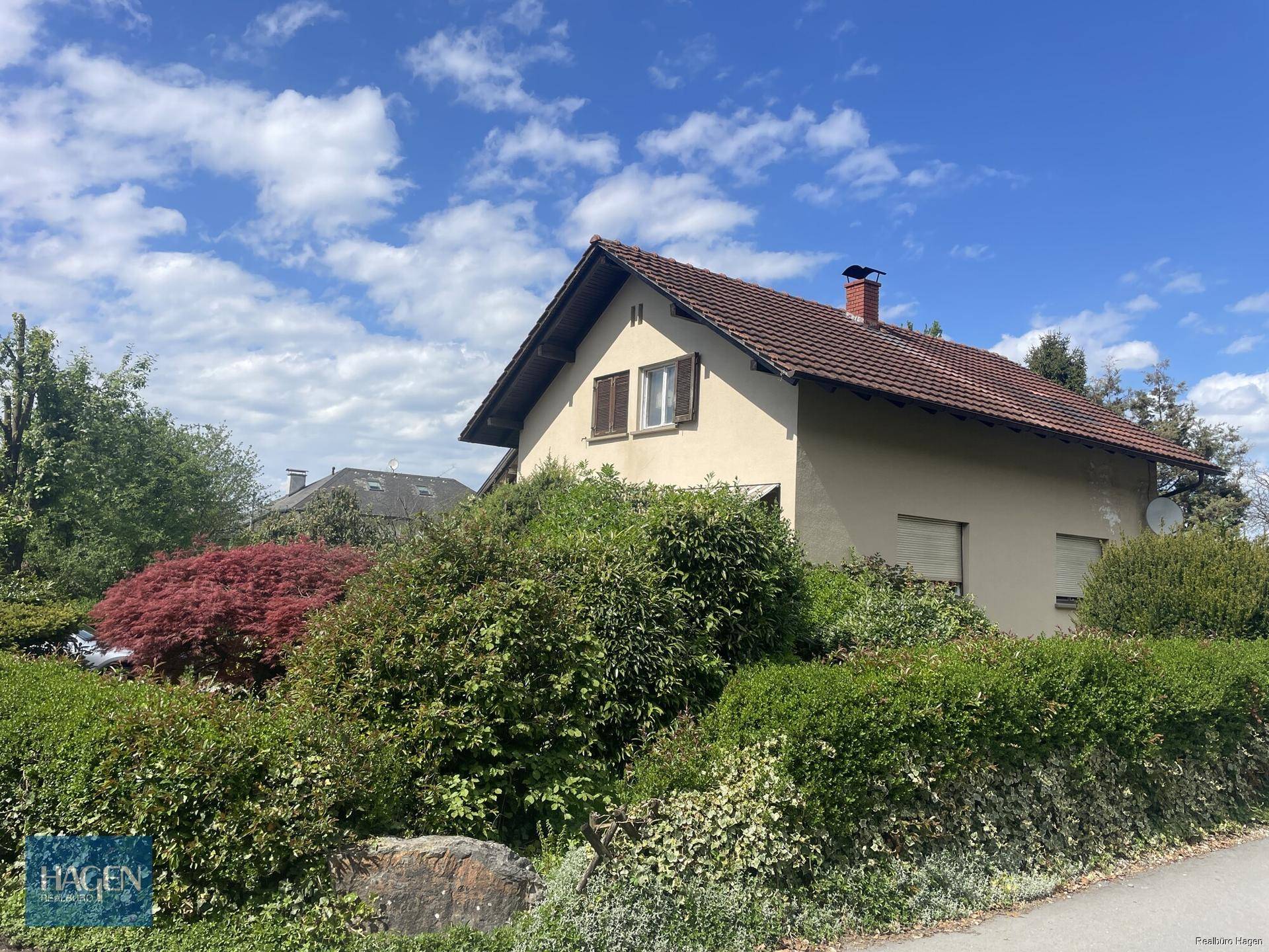 Ansicht des Hauses von der Straße mit beiger Fassade, braunen Fensterläden und dichtem Grün im Vordergrund.