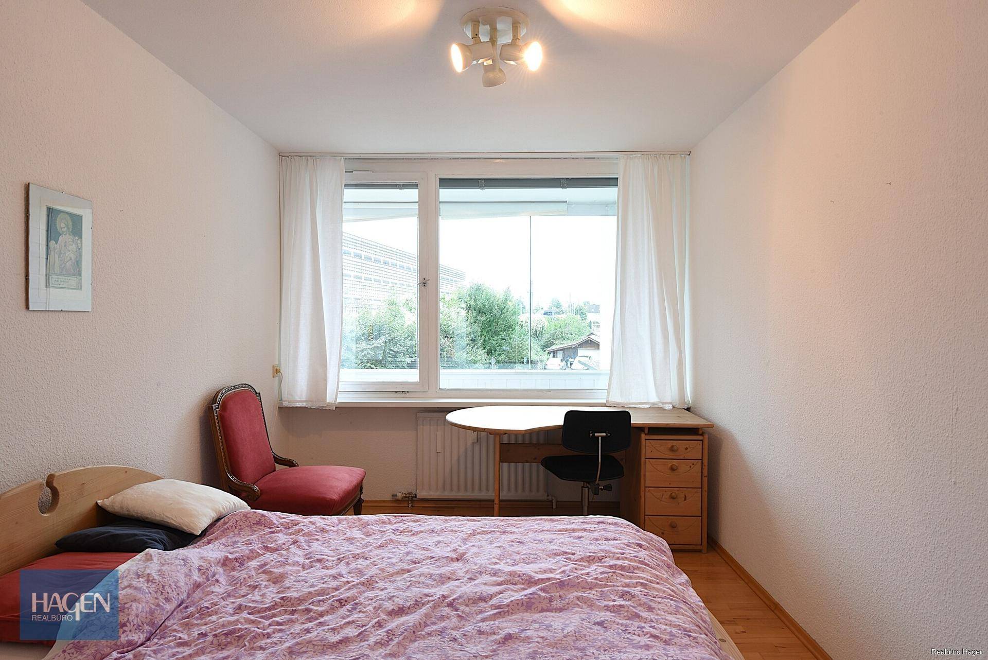 Schlafzimmer mit Holzboden, Bett und Schreibtisch, gut beleuchtet durch großes Fenster.