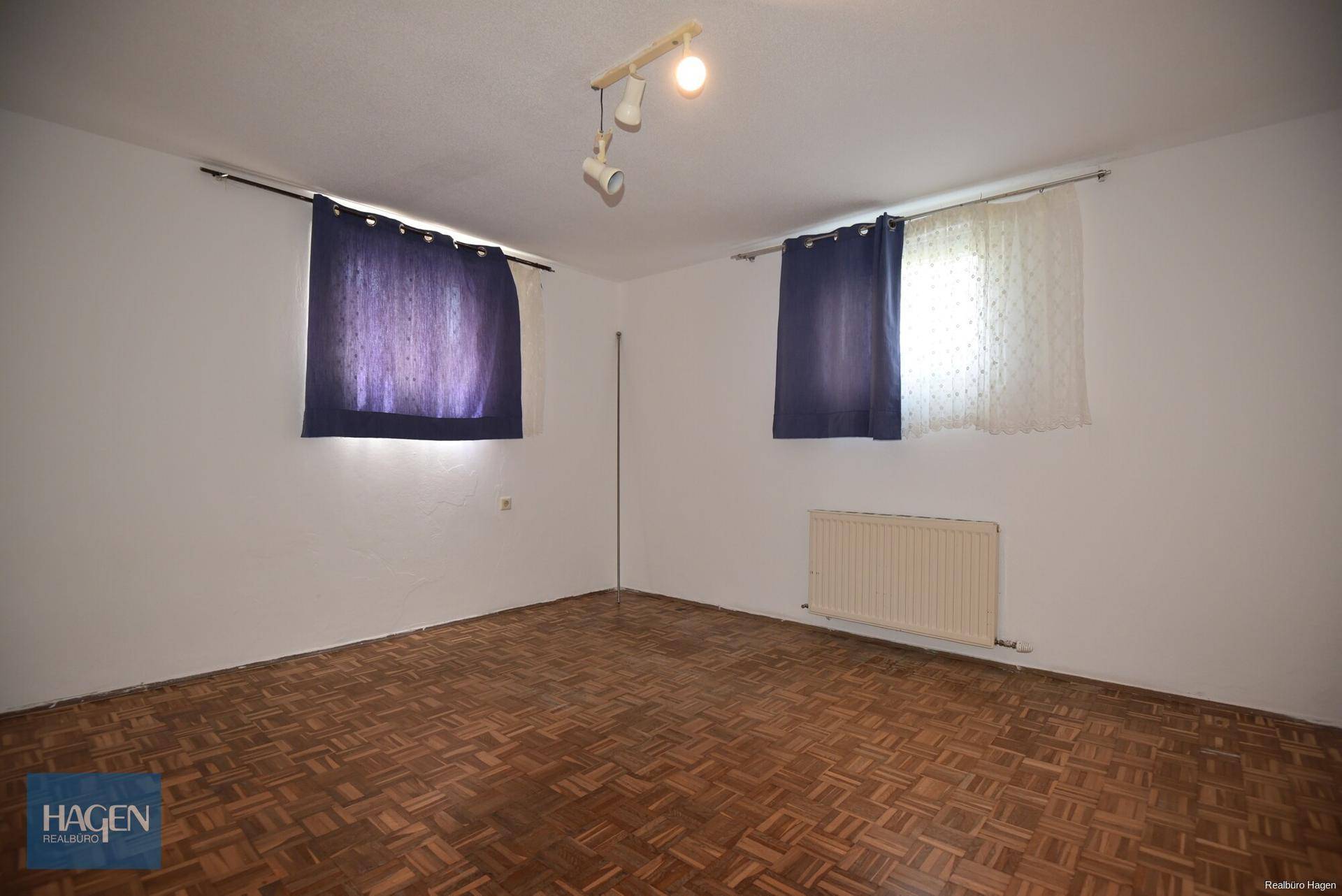 Helles Zimmer mit Parkettboden, zwei Fenstern und blauen Vorhängen.