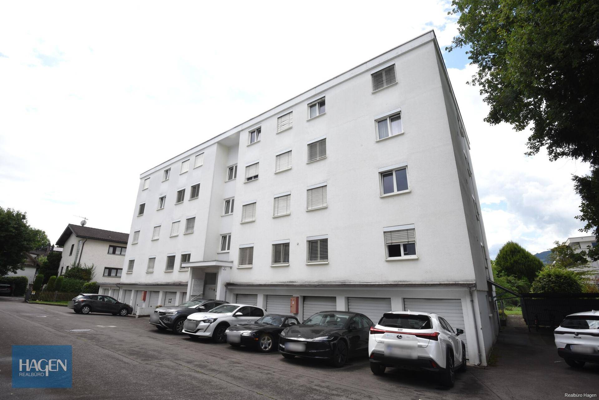 Mehrstöckiges Wohngebäude mit weißer Fassade und Parkplätzen vor dem Eingangsbereich.