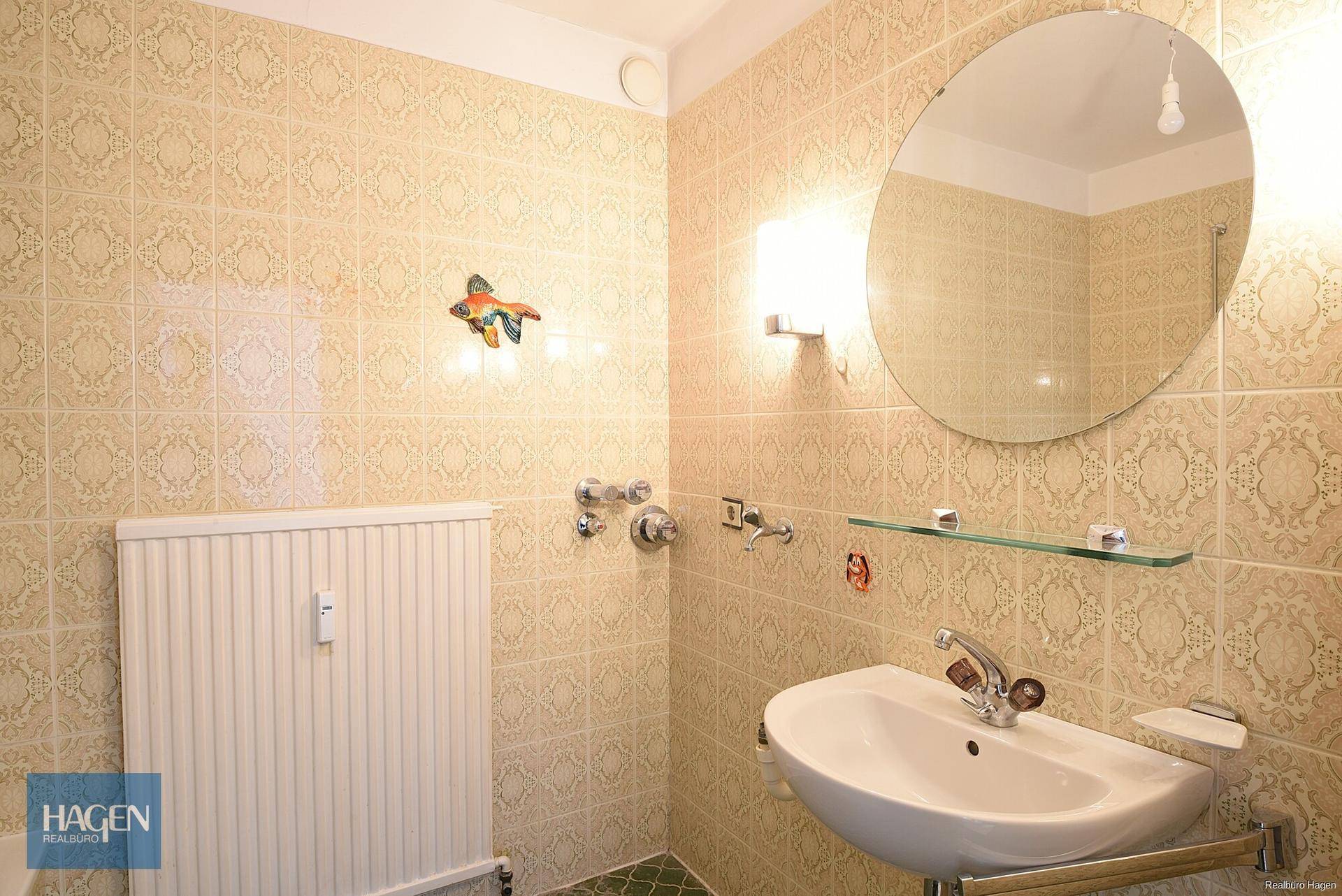Badezimmer mit gemusterten Fliesen, Waschbecken und rundem Spiegel.
