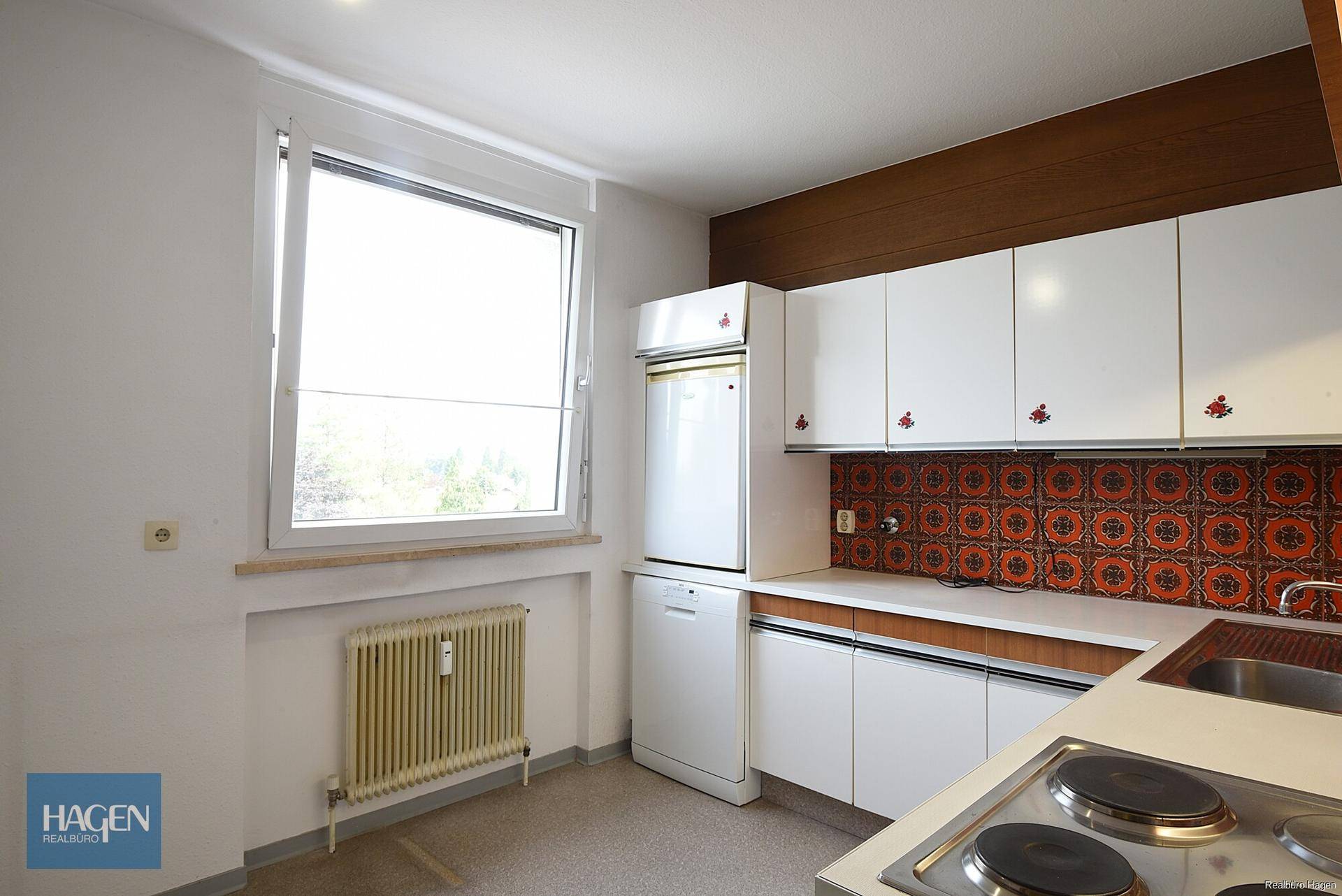 Küche mit Fenster, älteren Einbauschränken und gemusterten Fliesen an der Wand.