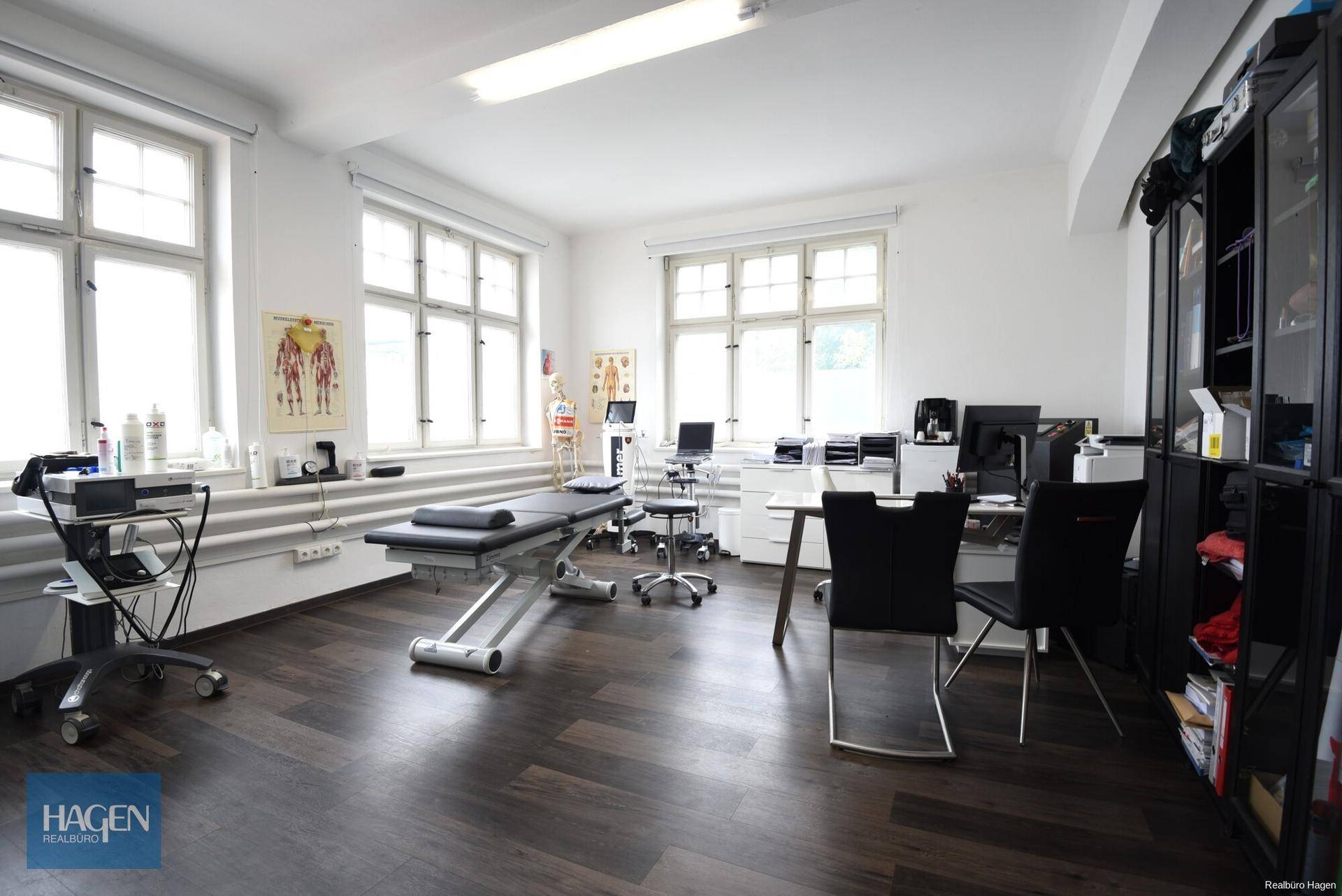 Therapieraum mit Behandlungsliege, Schreibtisch und medizinischen Wandtafeln.