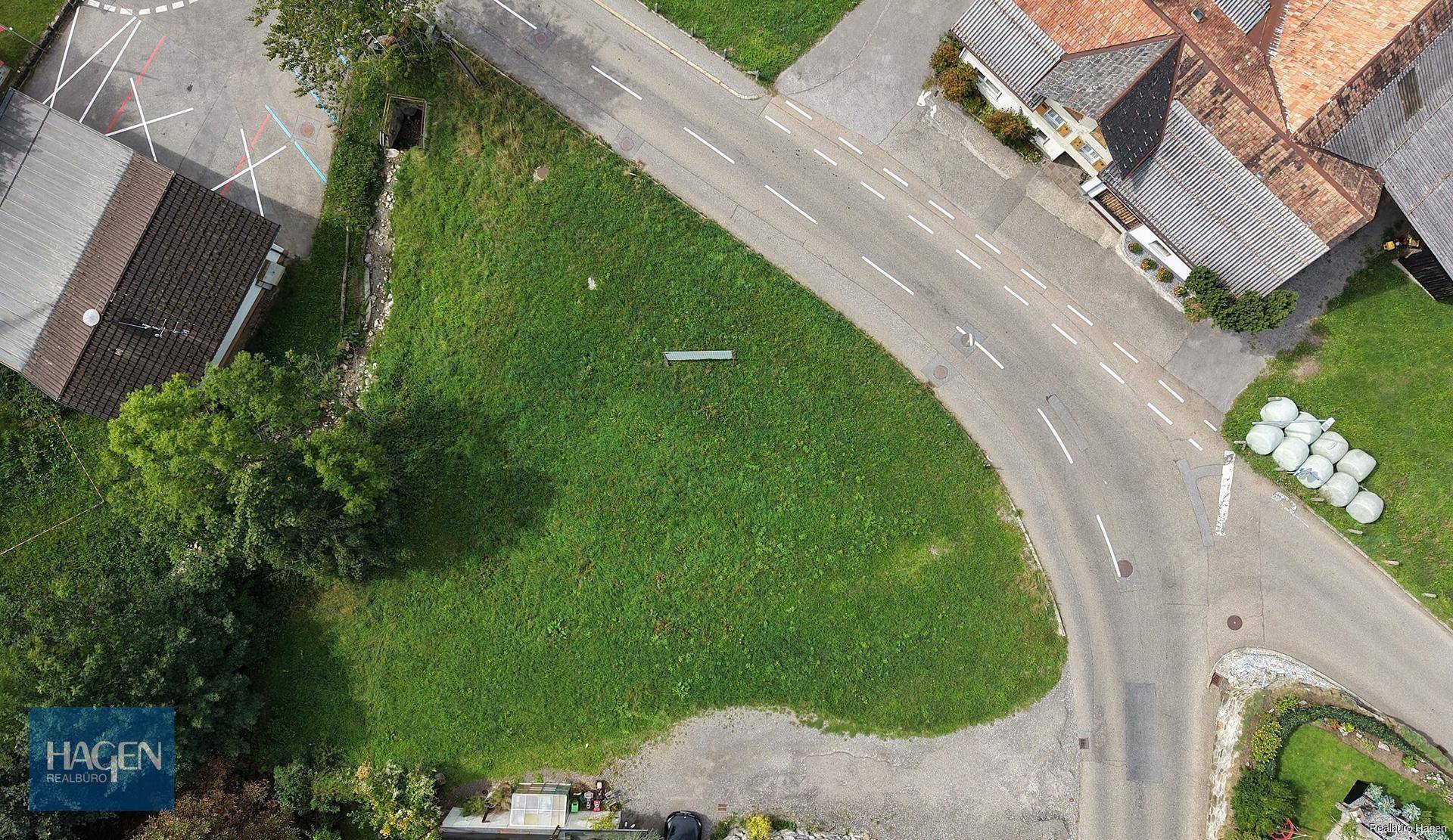 Senkrechte Luftaufnahme eines grünen Grundstücks an einer Straßenkreuzung mit angrenzenden Gebäuden.