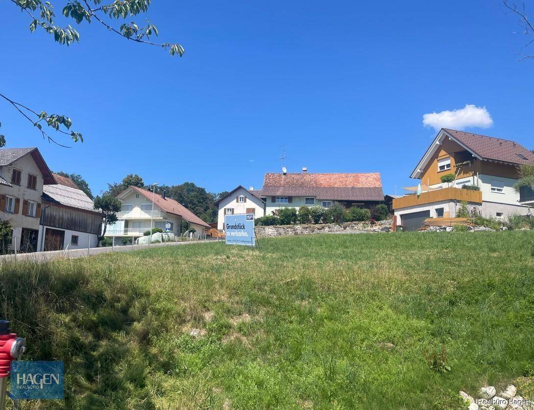 Blick auf ein sonniges Baugrundstück mit einem Schild und traditionellen Häusern im Hintergrund.