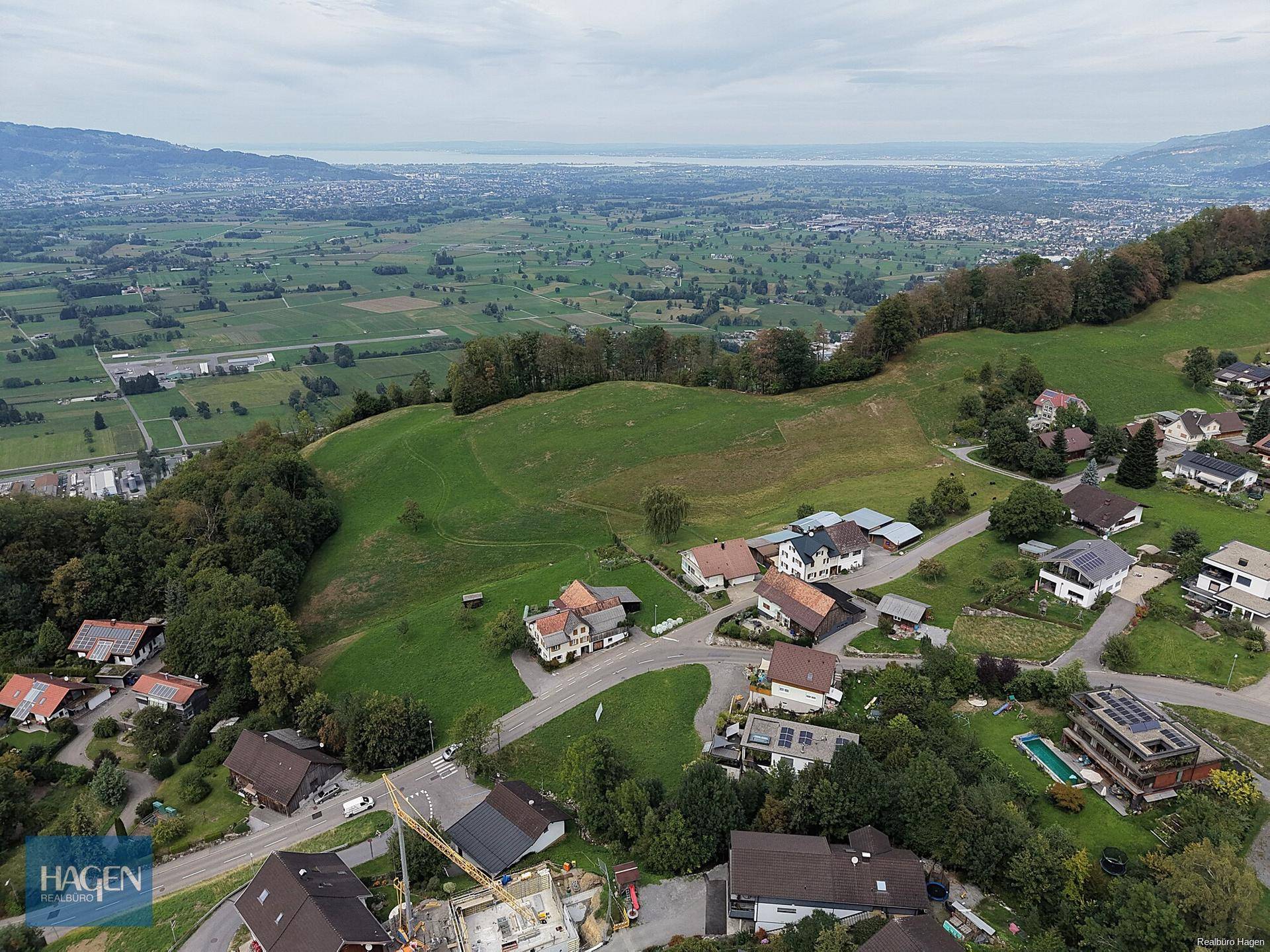 Weitläufige Luftaufnahme einer Wohnsiedlung in hügeliger Landschaft mit Blick auf einen See in der Ferne.