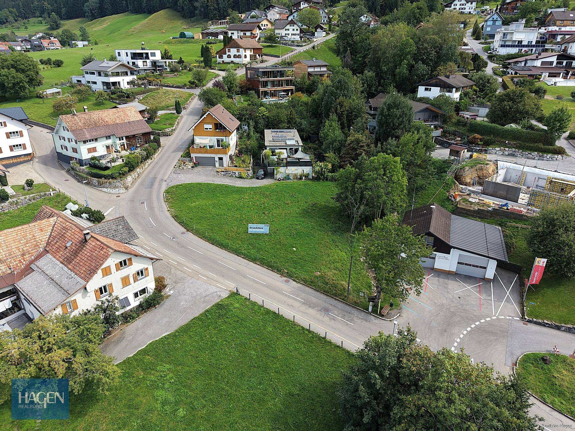 Luftaufnahme eines Baugrundstücks umgeben von Wohnhäusern und grünen Hügeln.