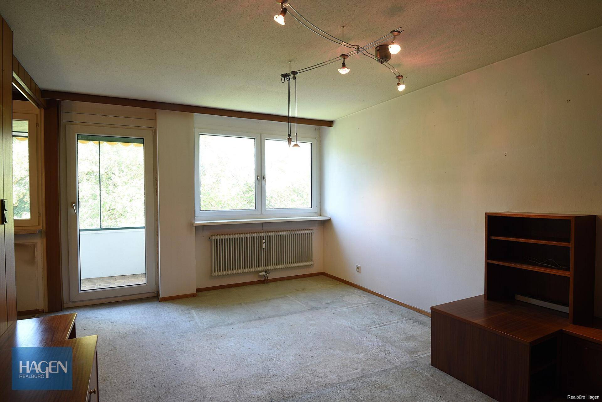 Helles Wohnzimmer mit Teppichboden, großem Fenster und Zugang zu einem Balkon.