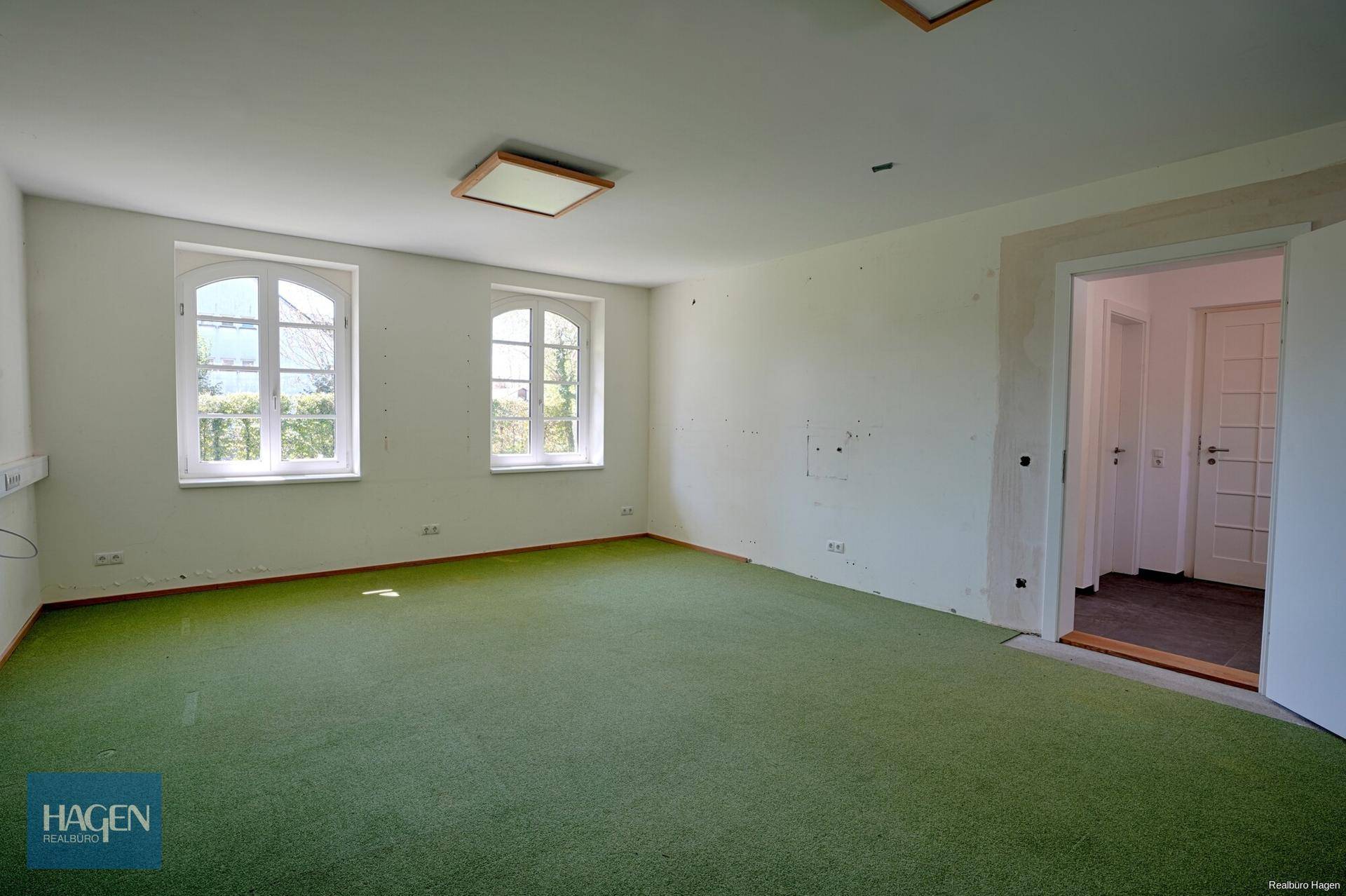 Geräumiges Zimmer mit grünem Teppichboden und zwei hohen Fenstern, bereit zur individuellen Gestaltung.