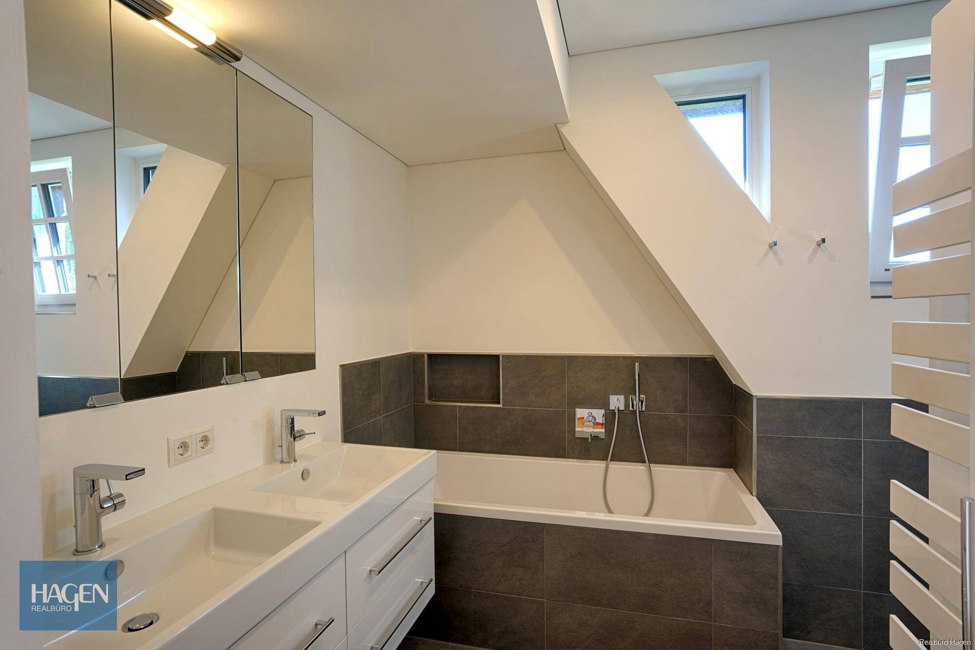 Modernes Badezimmer mit Doppelwaschbecken, Badewanne und grauen Fliesen unter Dachschräge.