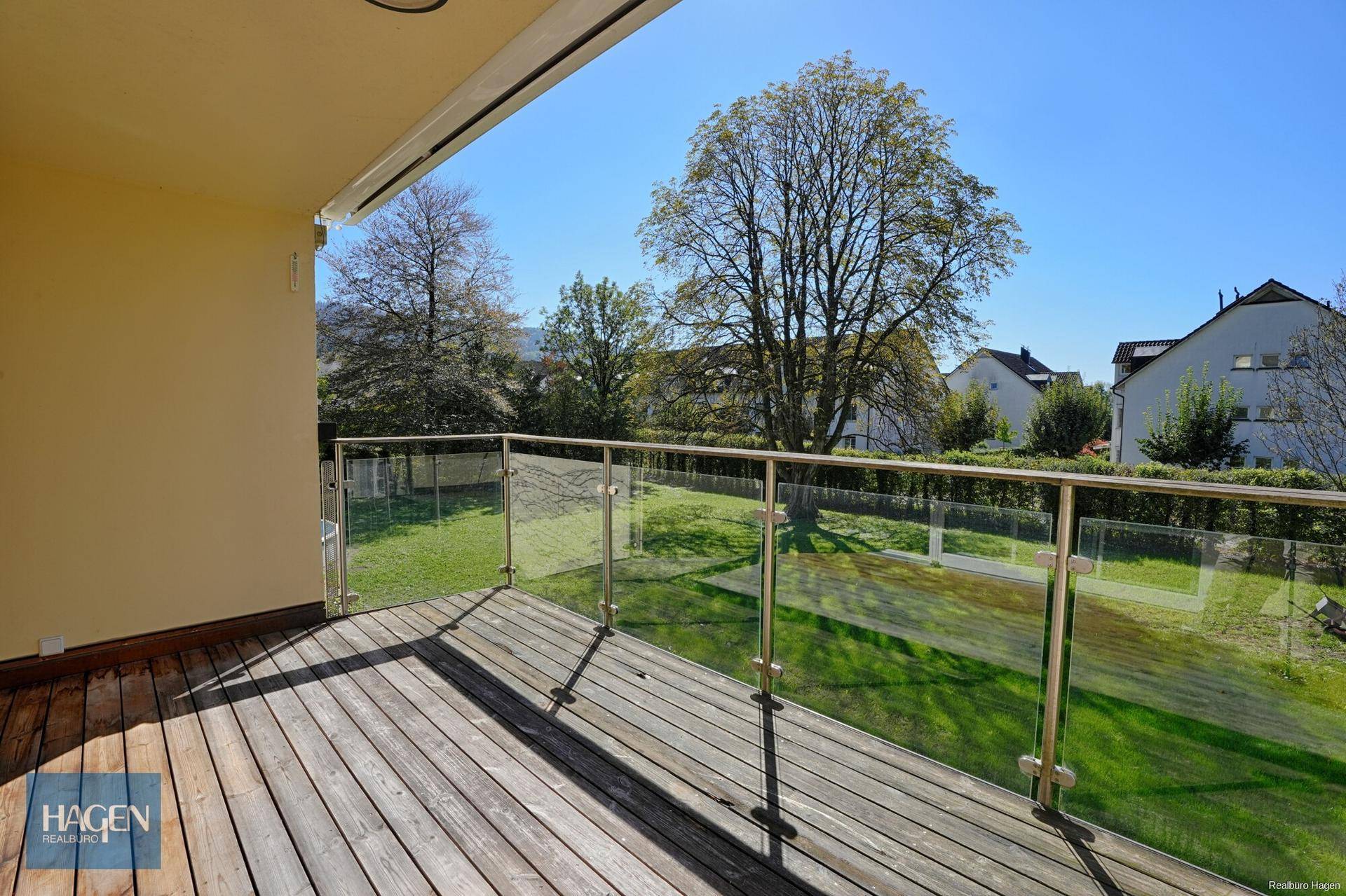 Geräumiger Balkon mit Holzdeck und Glasgeländer, bietet einen weiten Blick ins Grüne.