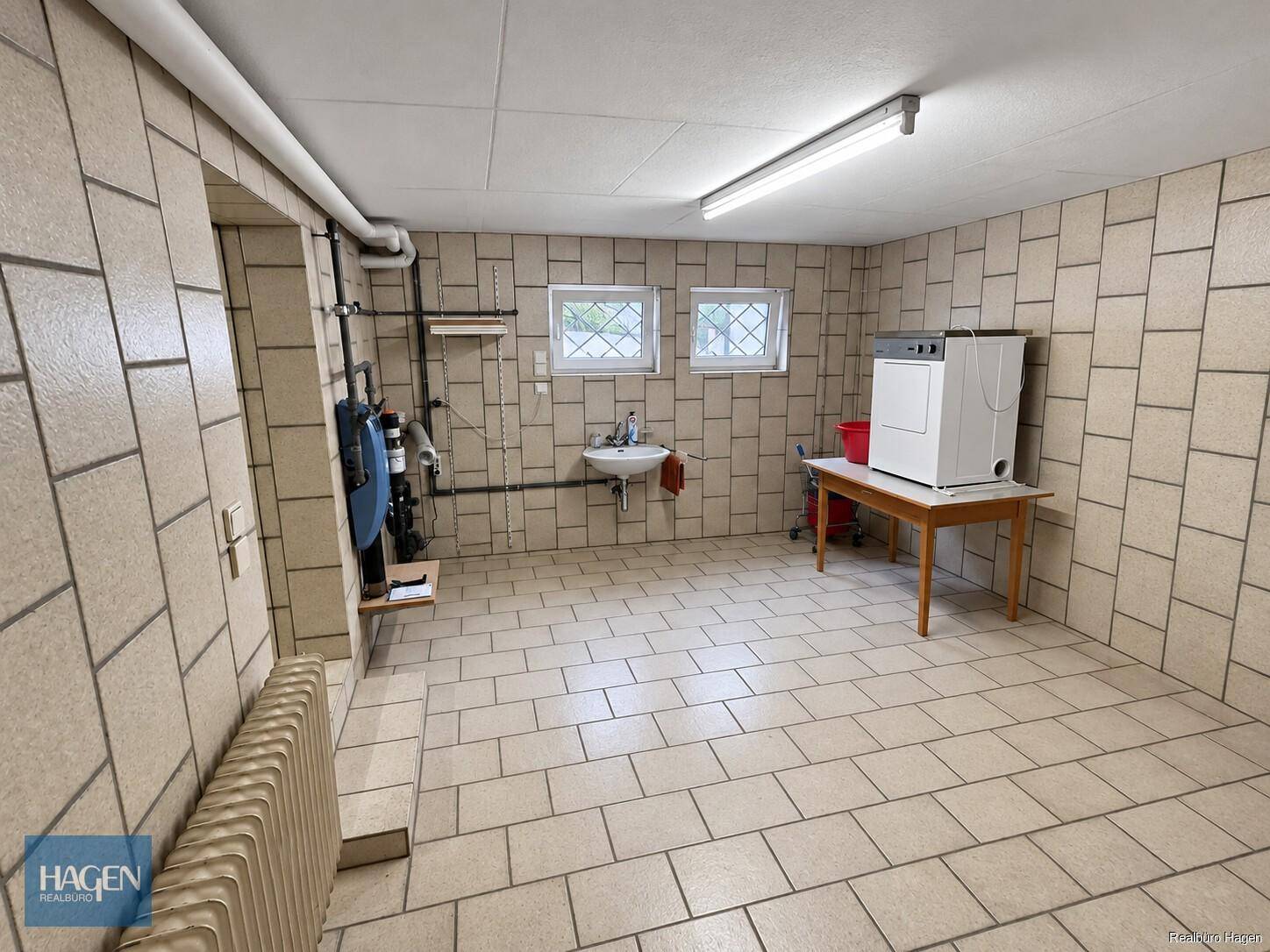 Heller Hauswirtschaftsraum mit Fliesenboden, Waschmaschine, Trockner und kleinem Waschbecken.