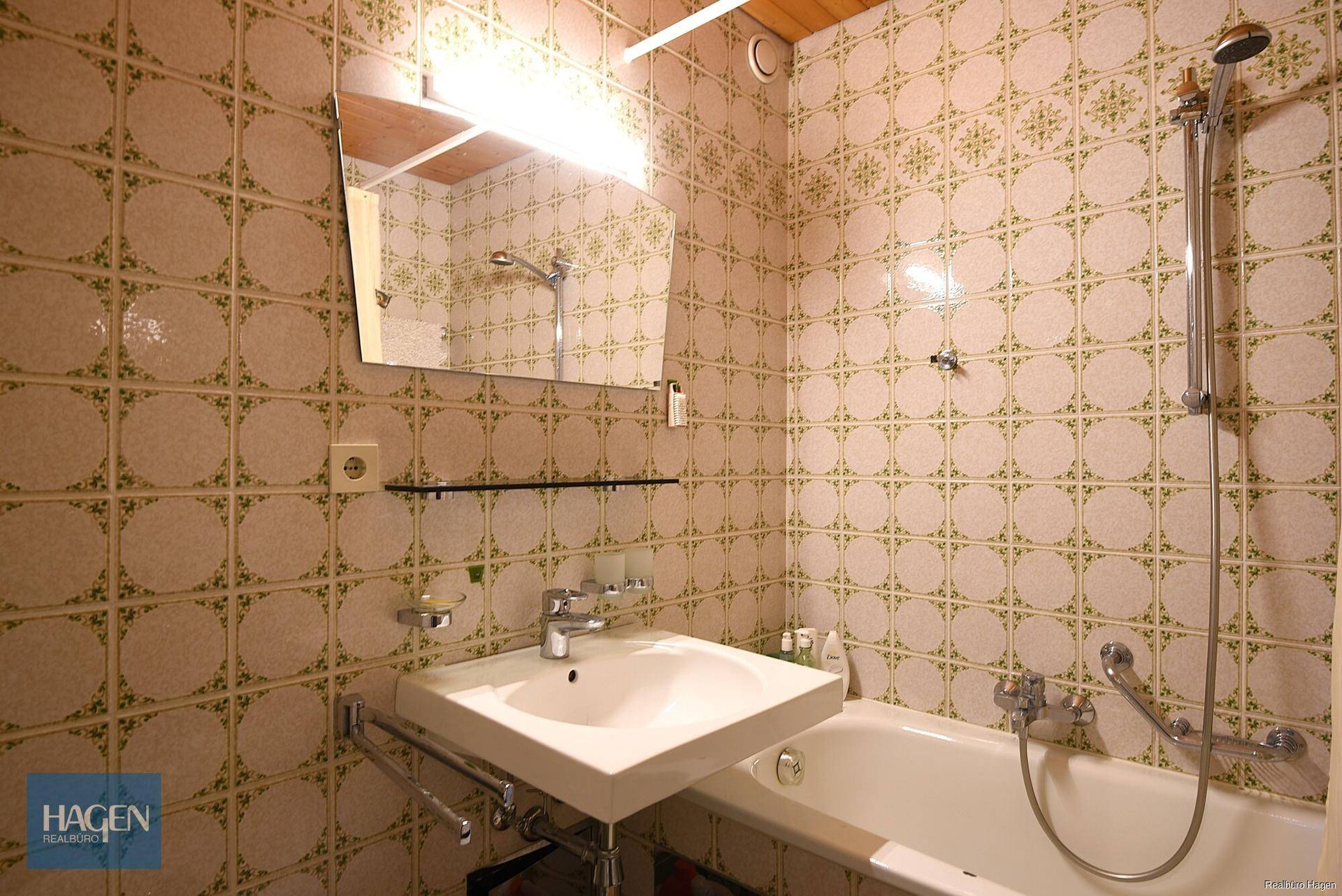 Badezimmer mit gemusterten Fliesen, Waschbecken, Spiegel und Badewanne mit Duschfunktion.