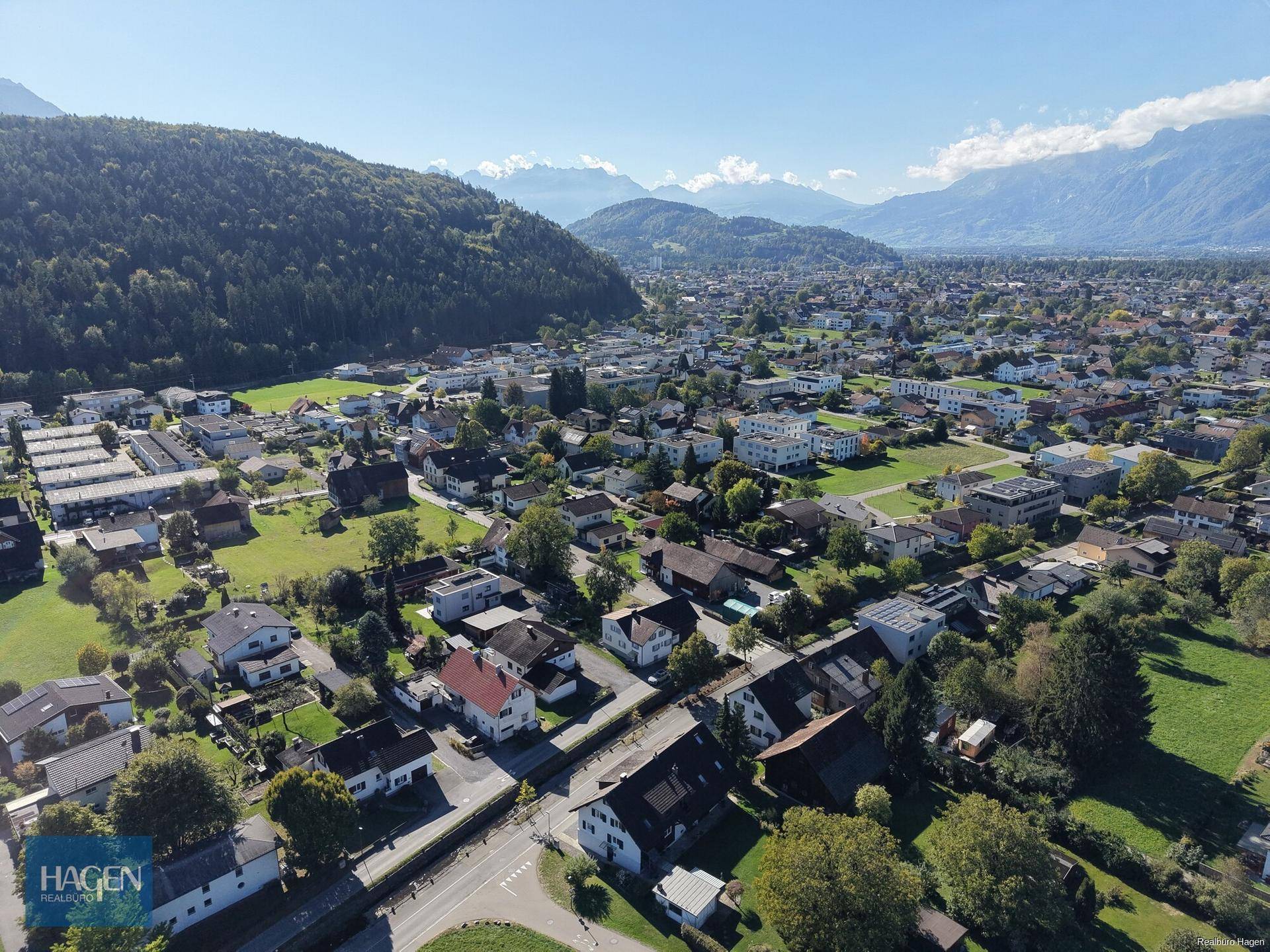 Weitläufige Luftaufnahme der Siedlung mit Häusern, Grünflächen und Bergen im Hintergrund.