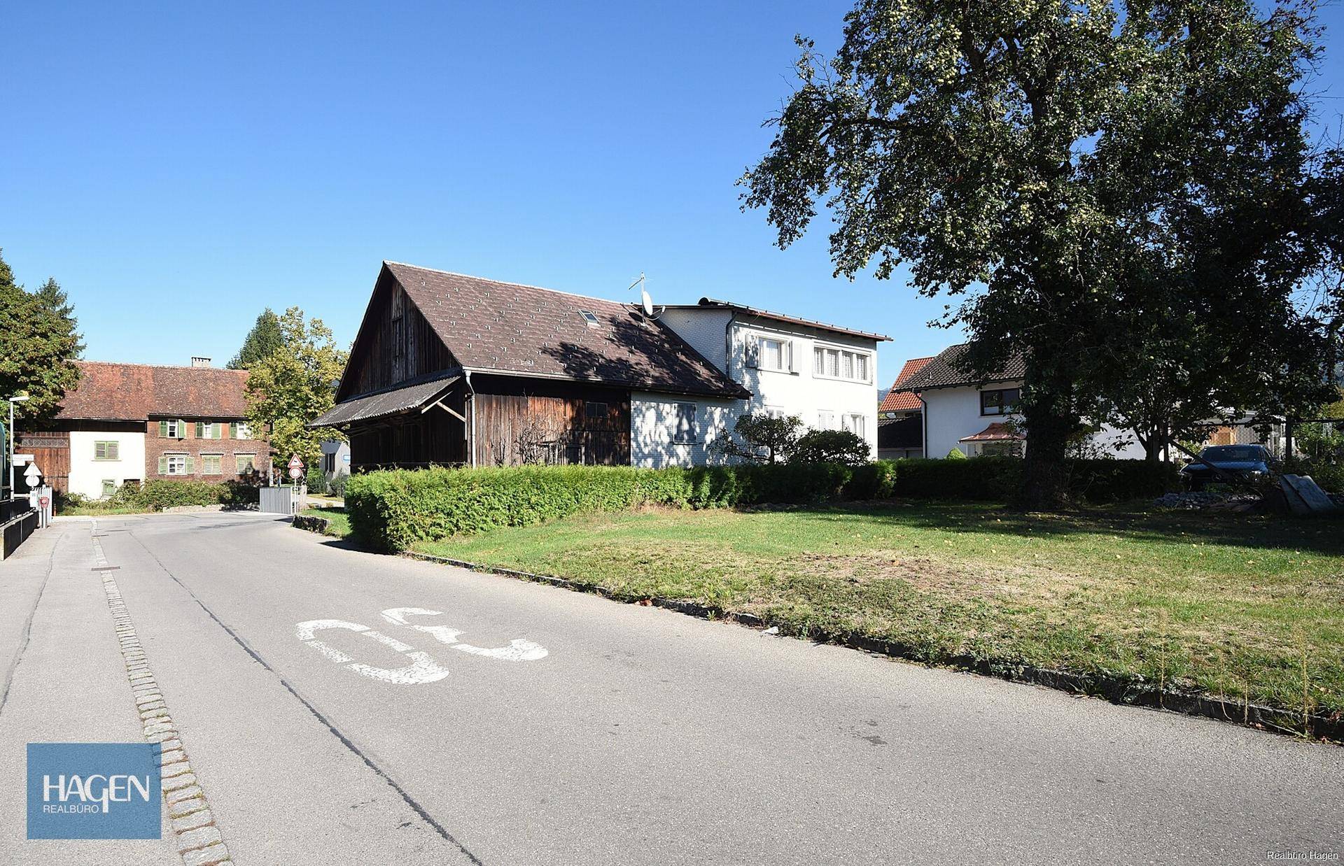 Ländliche Straße führt an einem traditionellen Holzhaus mit Satteldach vorbei.