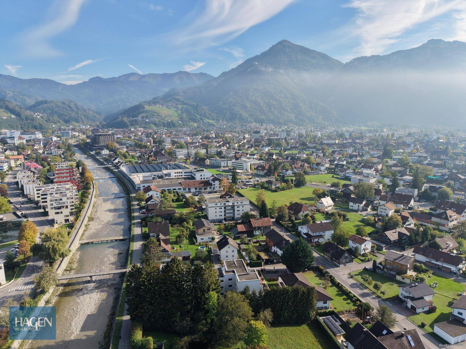 Luftaufnahme der Stadt mit Fluss, Wohngebieten und Bergen im Hintergrund.