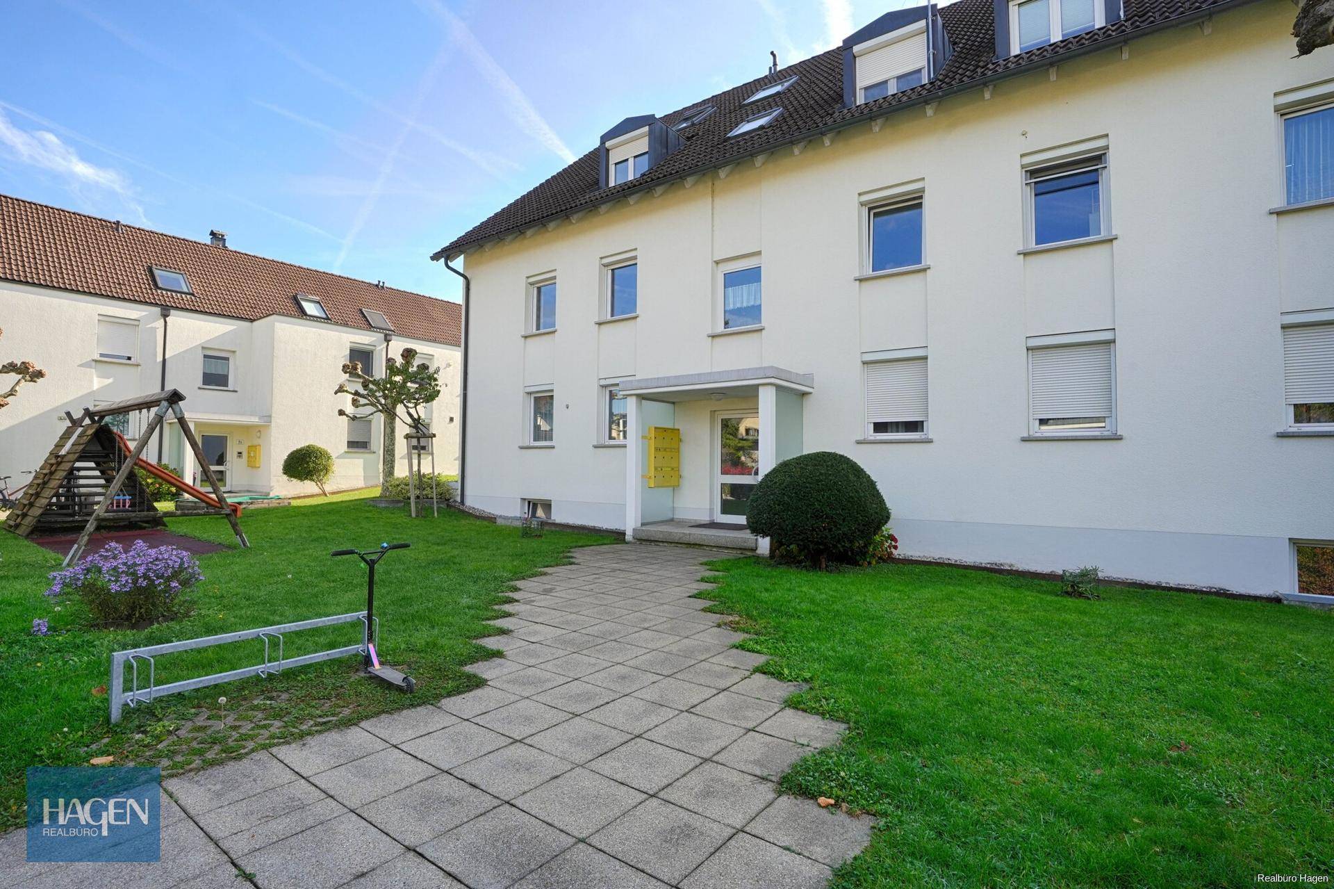 Mehrfamilienhaus mit weißer Fassade, umgeben von Grünflächen und einem Spielplatz.