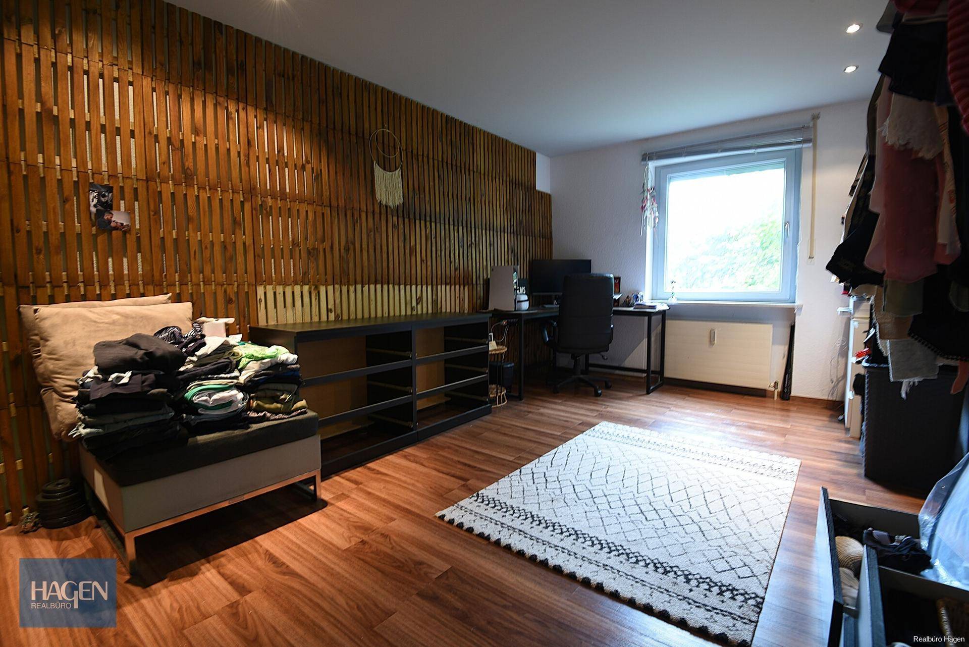 Schlafzimmer mit Holzwandverkleidung, Schreibtisch und Fenster mit Tageslicht.