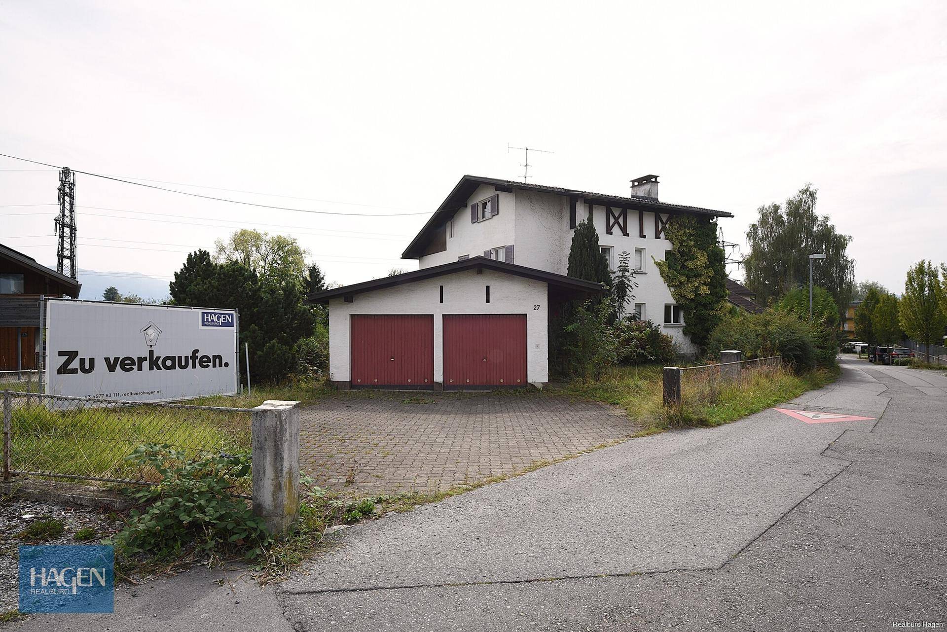Vorderansicht eines Wohnhauses mit Doppelgarage und einem 'Zu verkaufen'-Schild am Straßenrand.