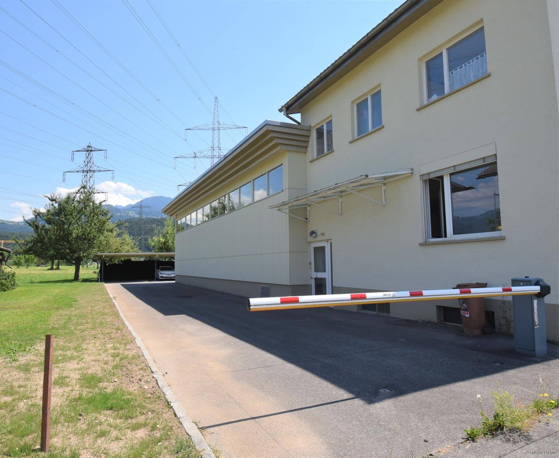 Seitenansicht des Gebäudes mit Zufahrt, Schranke und Strommasten im Hintergrund.