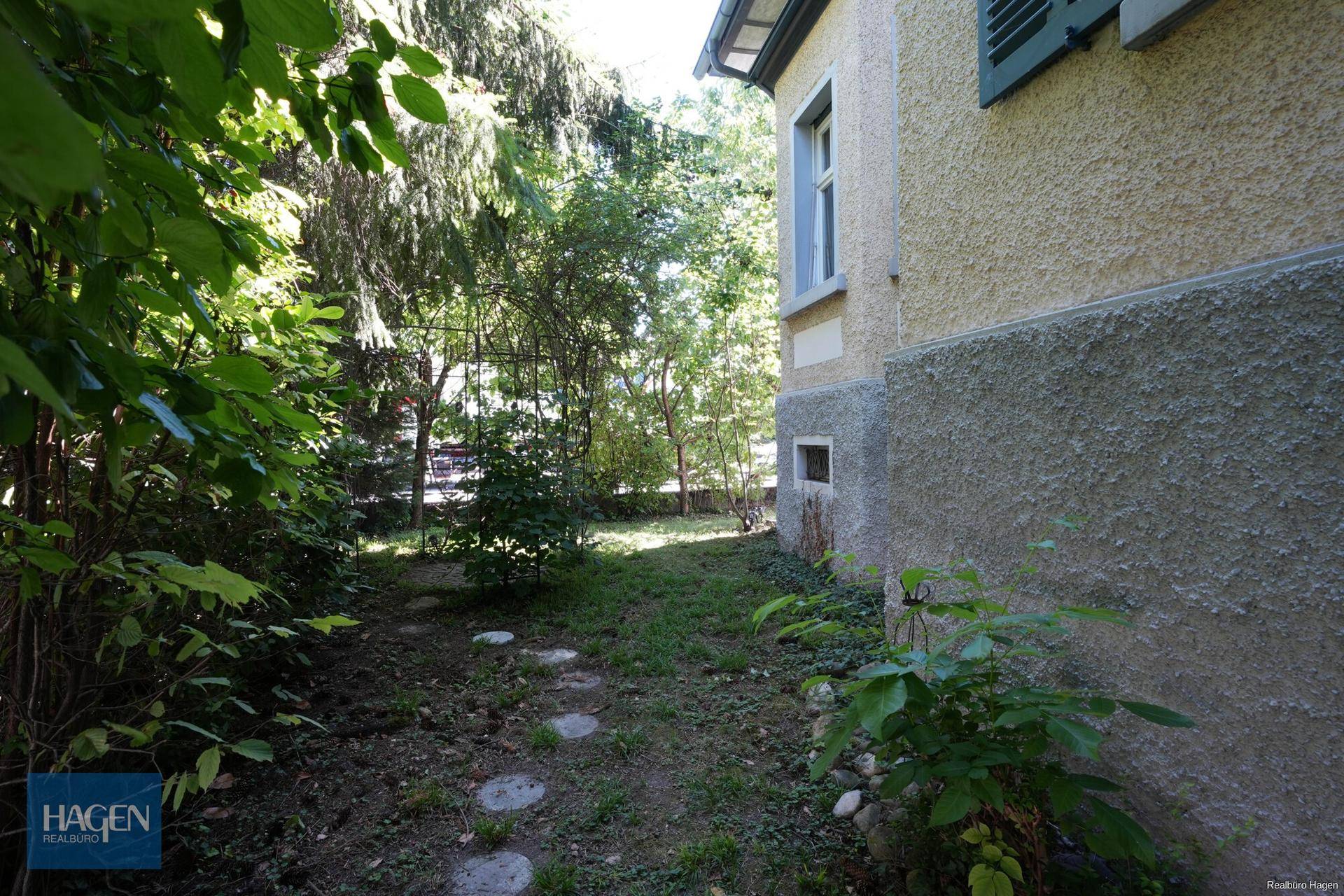Grüner Gartenbereich mit Bäumen, Sträuchern und einem kleinen Pfad neben dem Haus.
