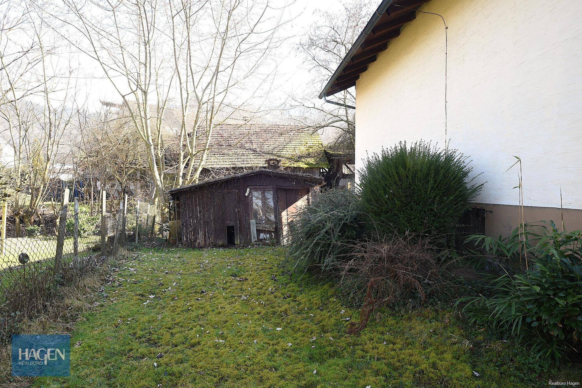Gartenbereich mit altem Holzschuppen und dichten Büschen, umgeben von einer natürlichen Landschaft.