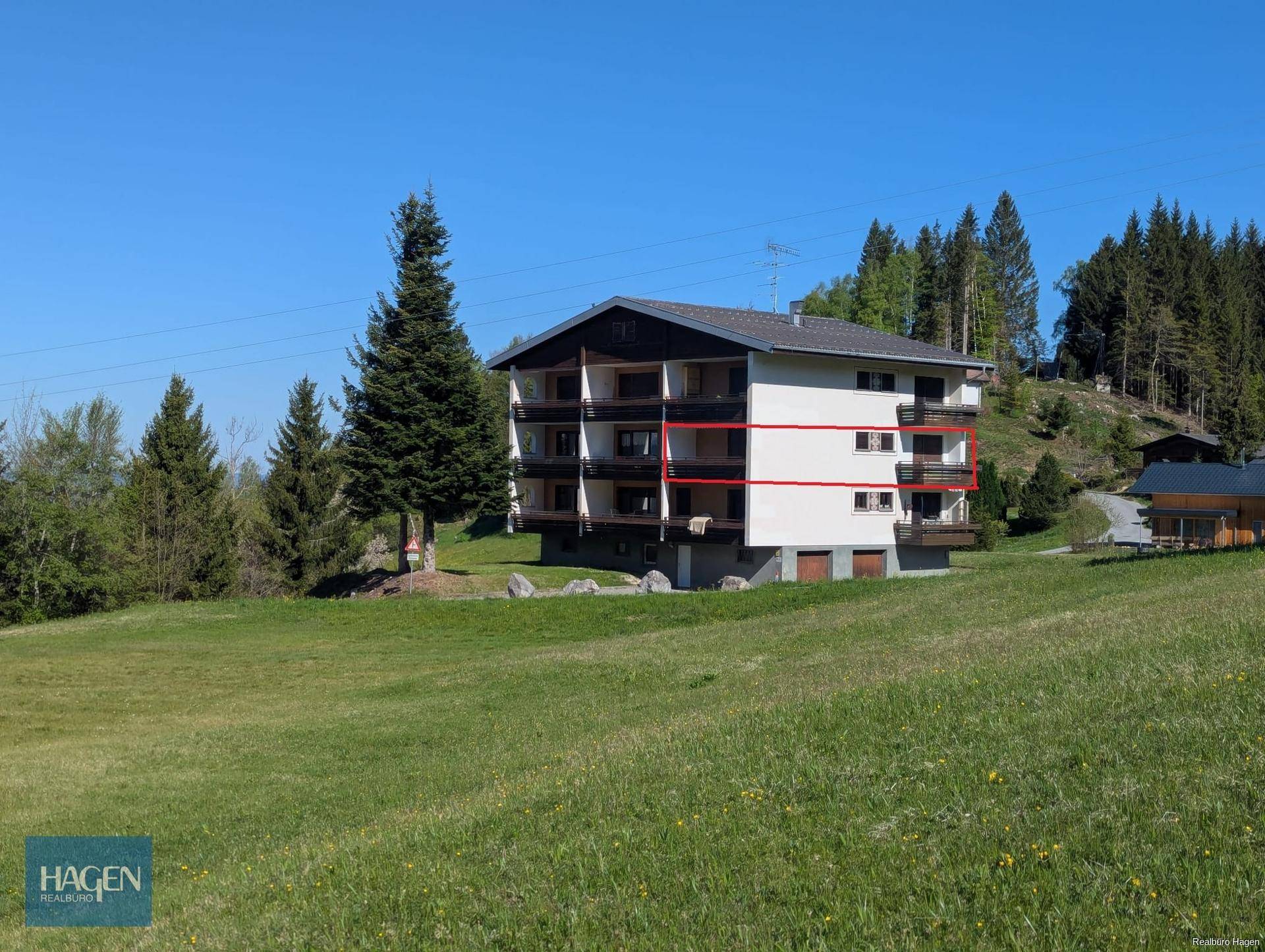 Ansicht des Mehrfamilienhauses mit Balkonen und der umliegenden grünen Wiesenlandschaft unter blauem Himmel.