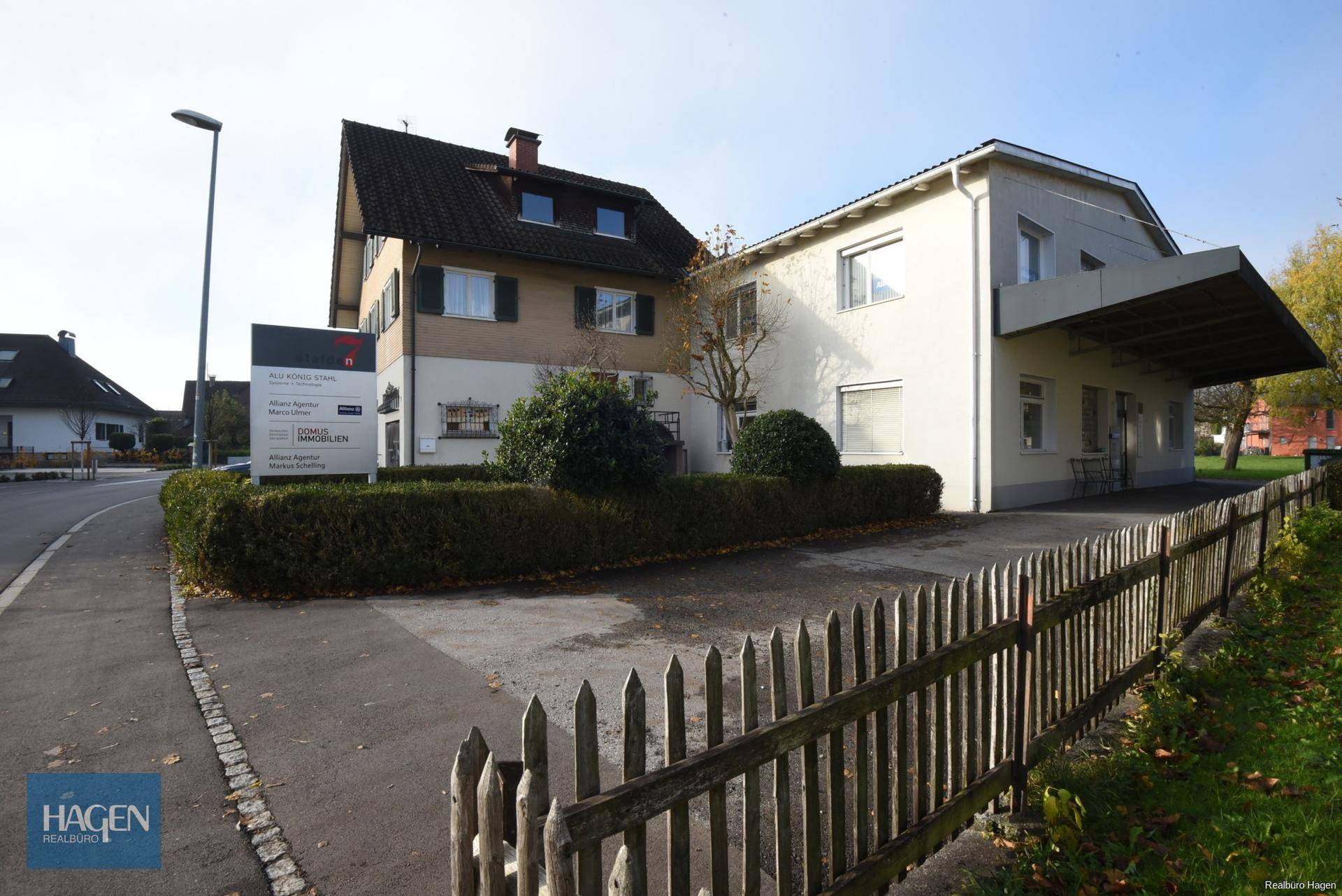 Ansicht des Gebäudes von der Straße mit einem Holzzaun und gepflegten Hecken im Vordergrund.