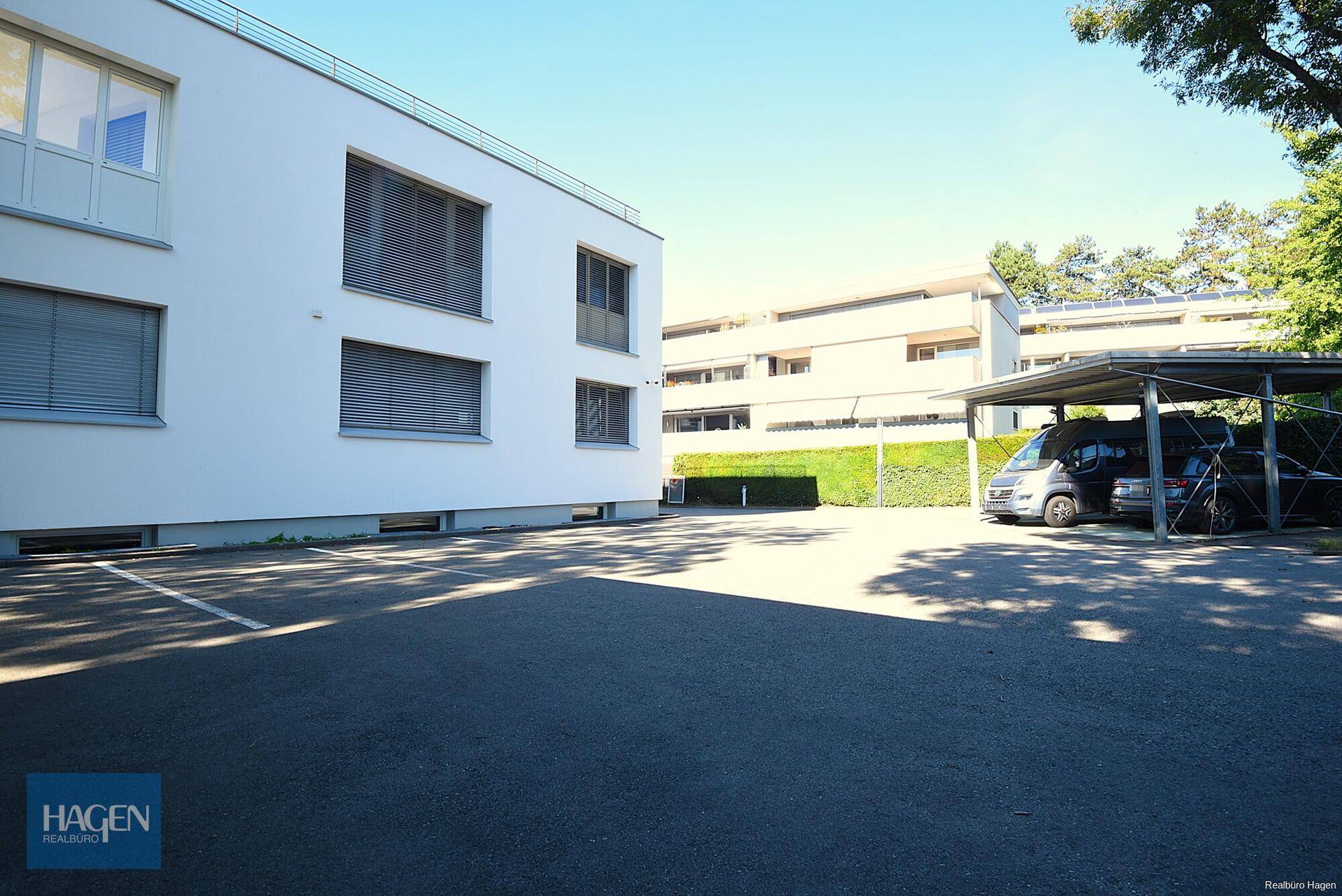 Außenansicht des Gebäudes mit Parkplätzen und einem Carport im Hintergrund.