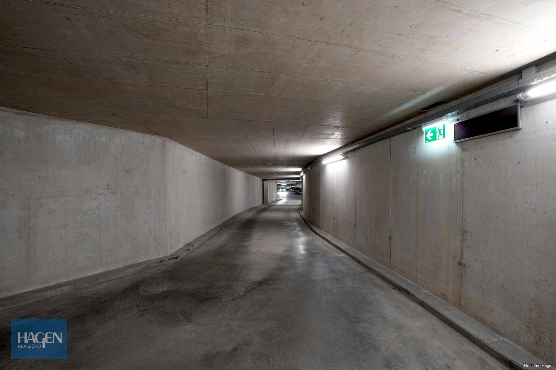 Tiefgarage mit Betonwänden und -decke, beleuchtet und mit Notausgangsschildern.