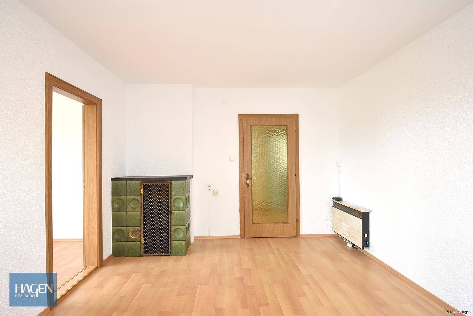 Geräumiges Wohnzimmer mit Laminatboden, weiß gestrichenen Wänden und einem traditionellen Kachelofen.