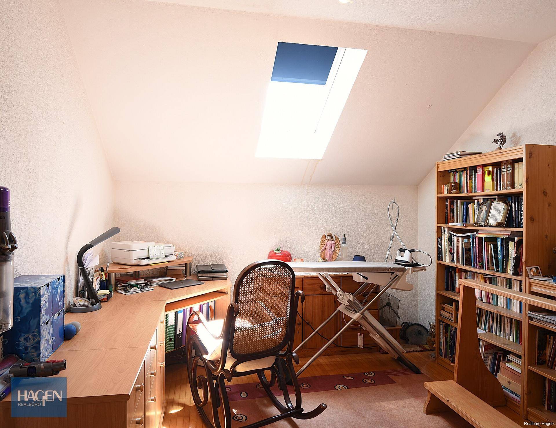 Helles Dachzimmer als Büro genutzt mit Schreibtisch, Bücherregal und Dachfenster.
