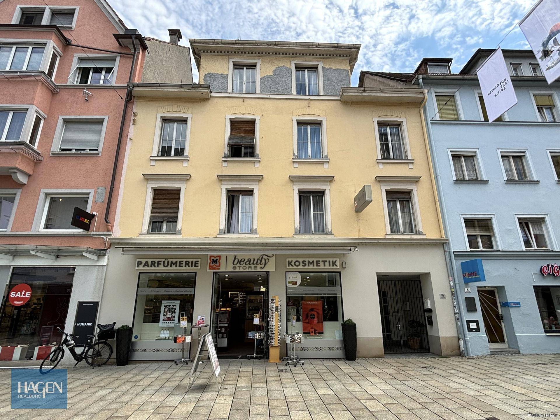 Gelbe Fassade eines Mehrfamilienhauses mit Geschäften im Erdgeschoss an einer belebten Straße.