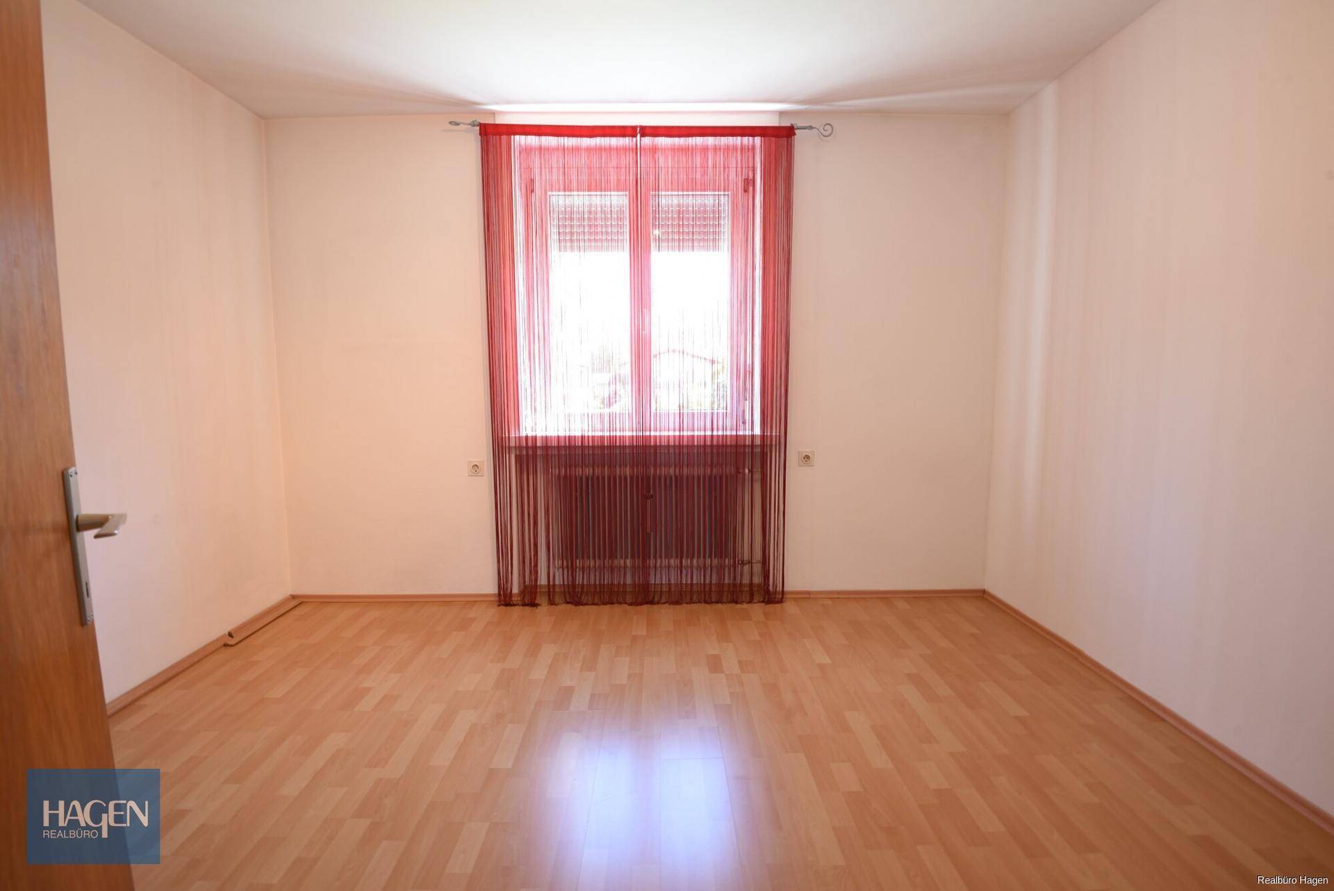 Helles Schlafzimmer mit Laminatboden und rotem Vorhang vor dem Fenster.