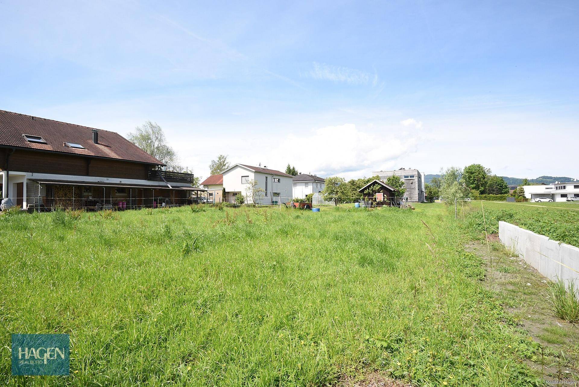 Weitläufiges grünes Grundstück mit hohem Gras und umliegenden Wohnhäusern unter blauem Himmel.