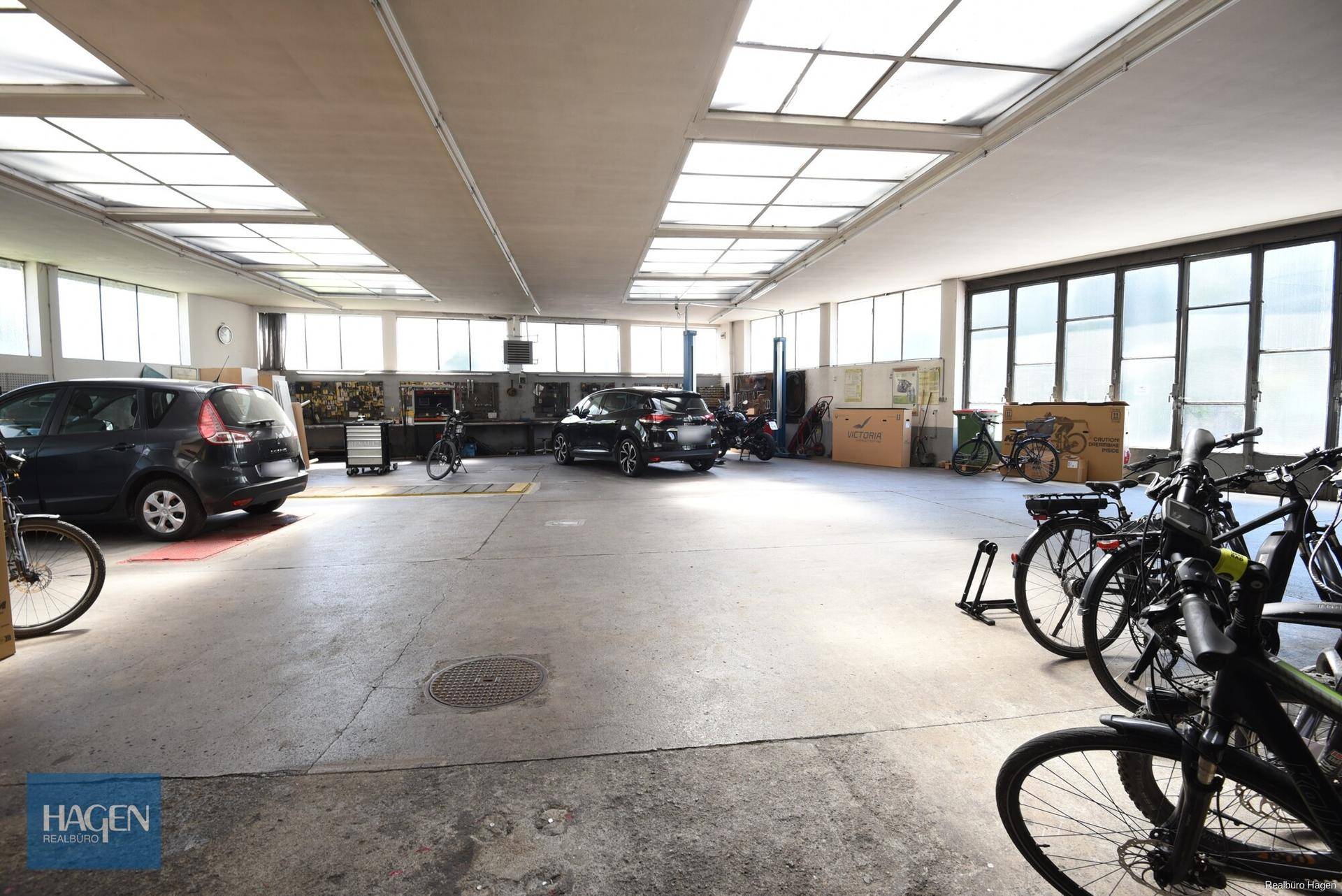 Großer Werkstatt- und Garagenbereich mit mehreren Fahrzeugen und Fahrrädern, Betonboden und Oberlichtern.