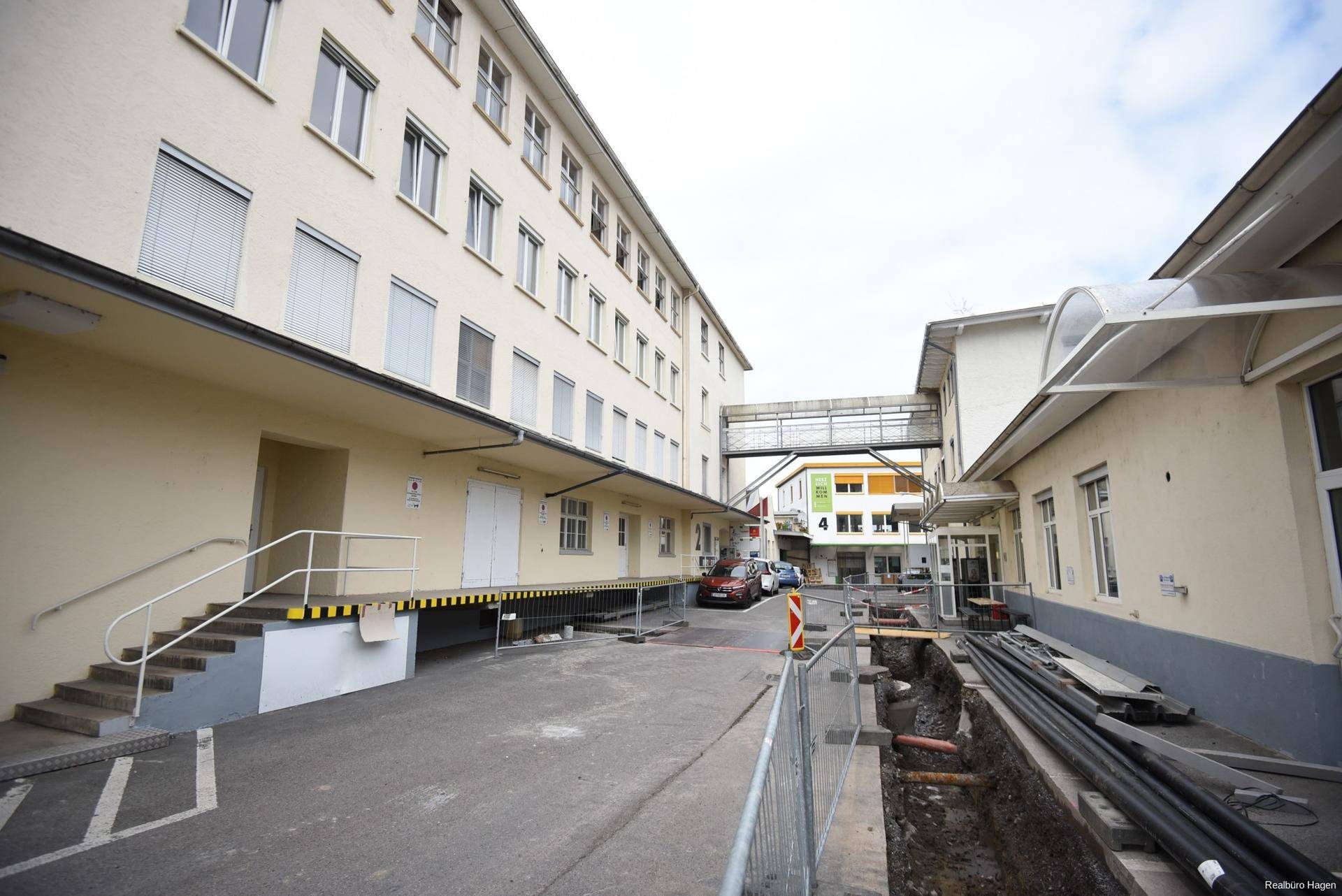 Außenansicht eines Geschäftsgebäudes mit Laderampen, einer Fußgängerbrücke und Bauarbeiten im Vordergrund.