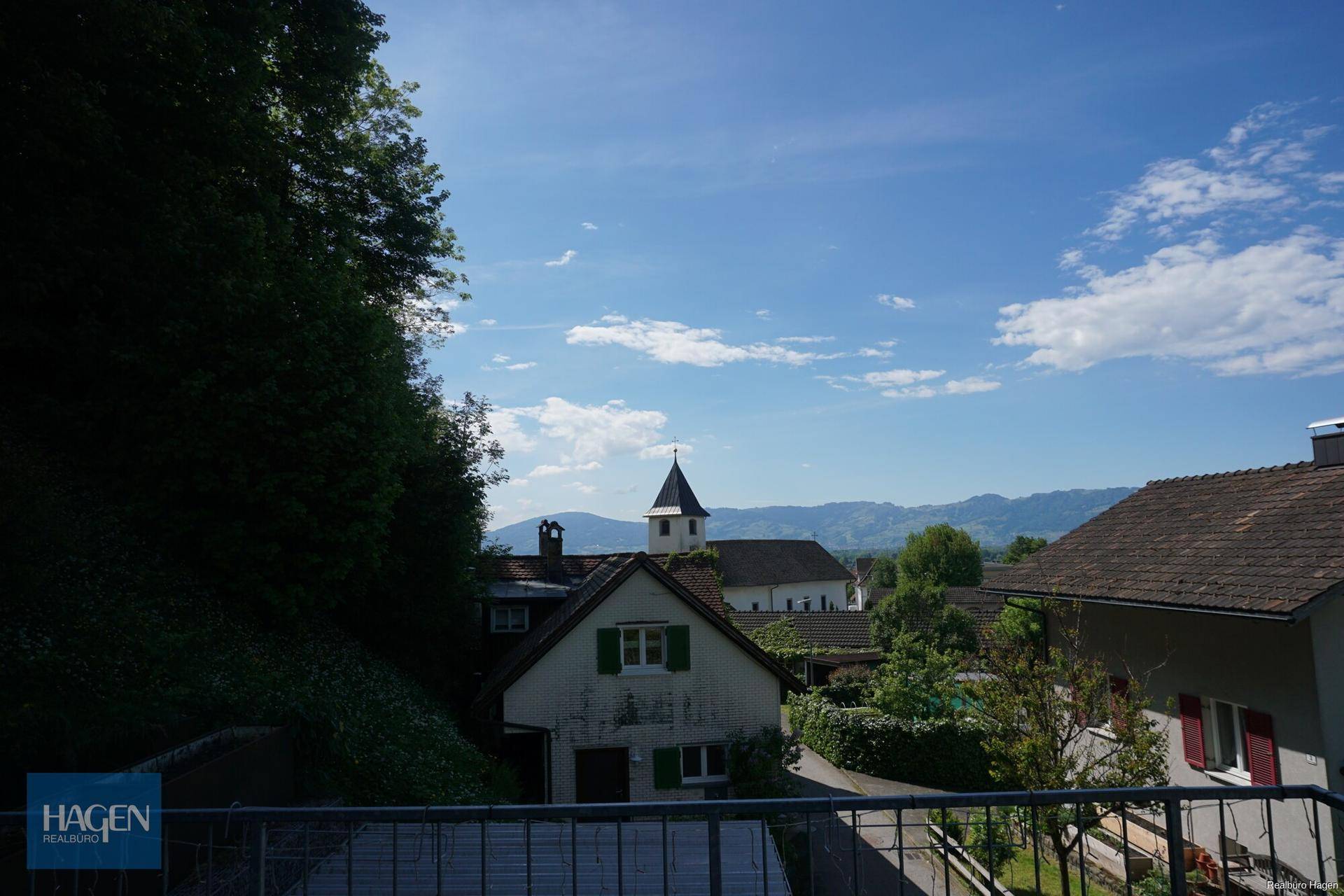 Panoramablick von einem Balkon auf ein Dorf mit Kirchturm und die umliegende Berglandschaft.
