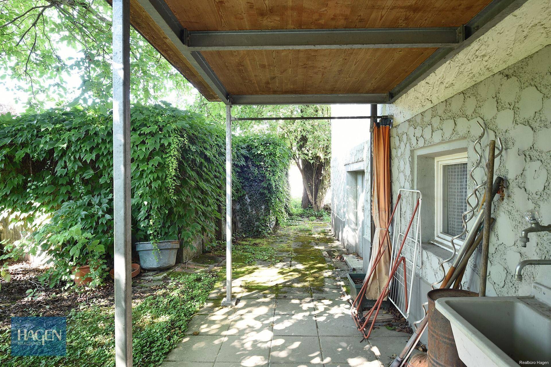 Überdachter Außenbereich mit Betonboden, Spülbecken und Blick auf den verwilderten Garten.