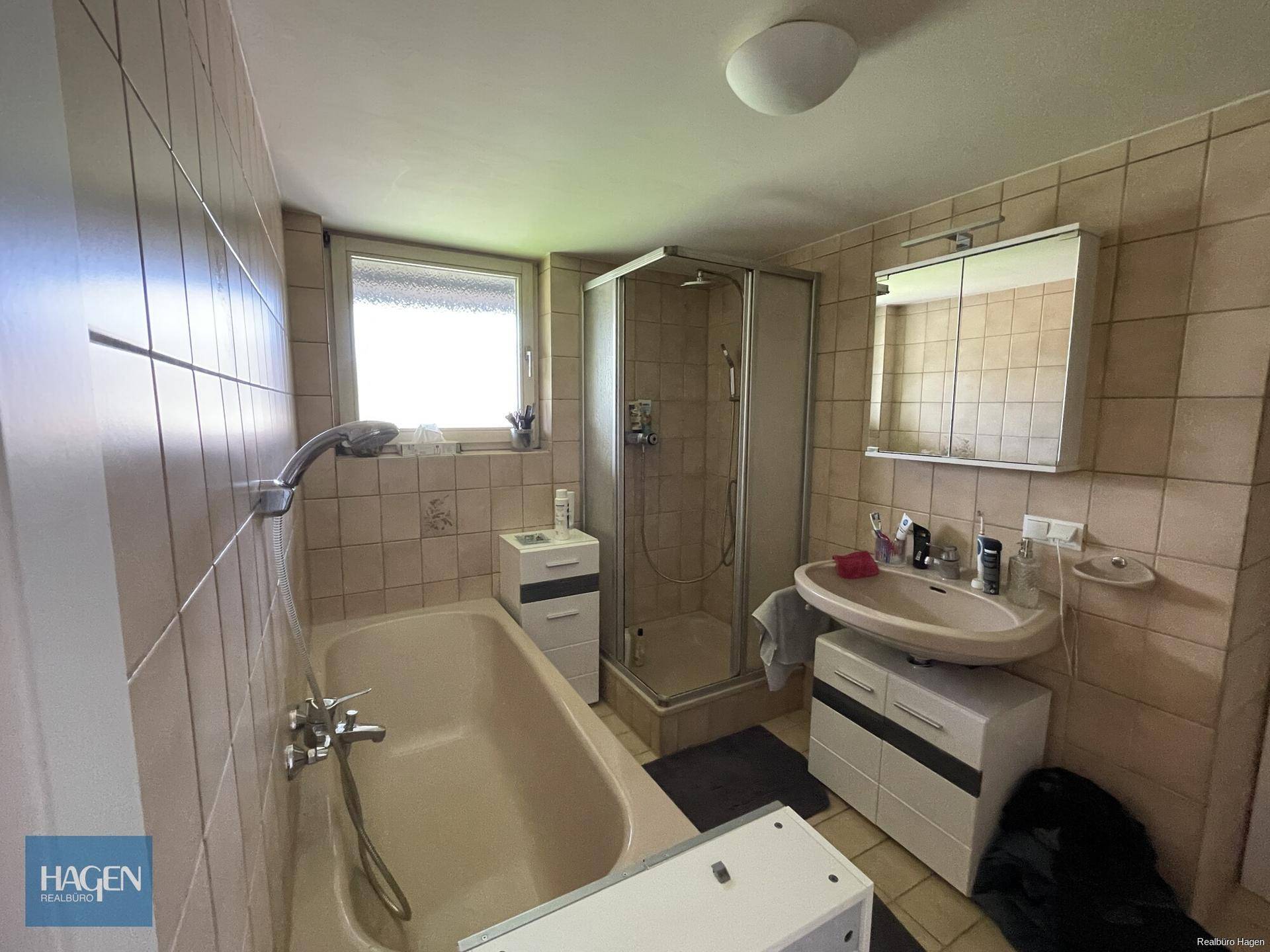 Funktionales Badezimmer mit beigen Fliesen, Badewanne, Duschkabine und Fenster.