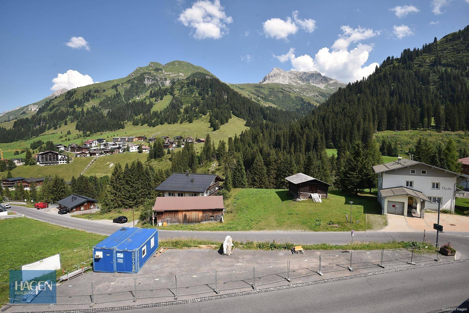 Sommerliche Ansicht von Häusern in einer Berglandschaft mit grünen Wiesen und Wäldern.