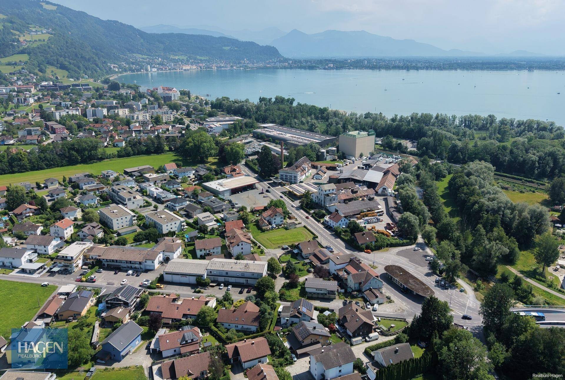 Weitläufige Luftaufnahme einer Stadt am See, umgeben von grünen Hügeln und Bergen unter blauem Himmel.